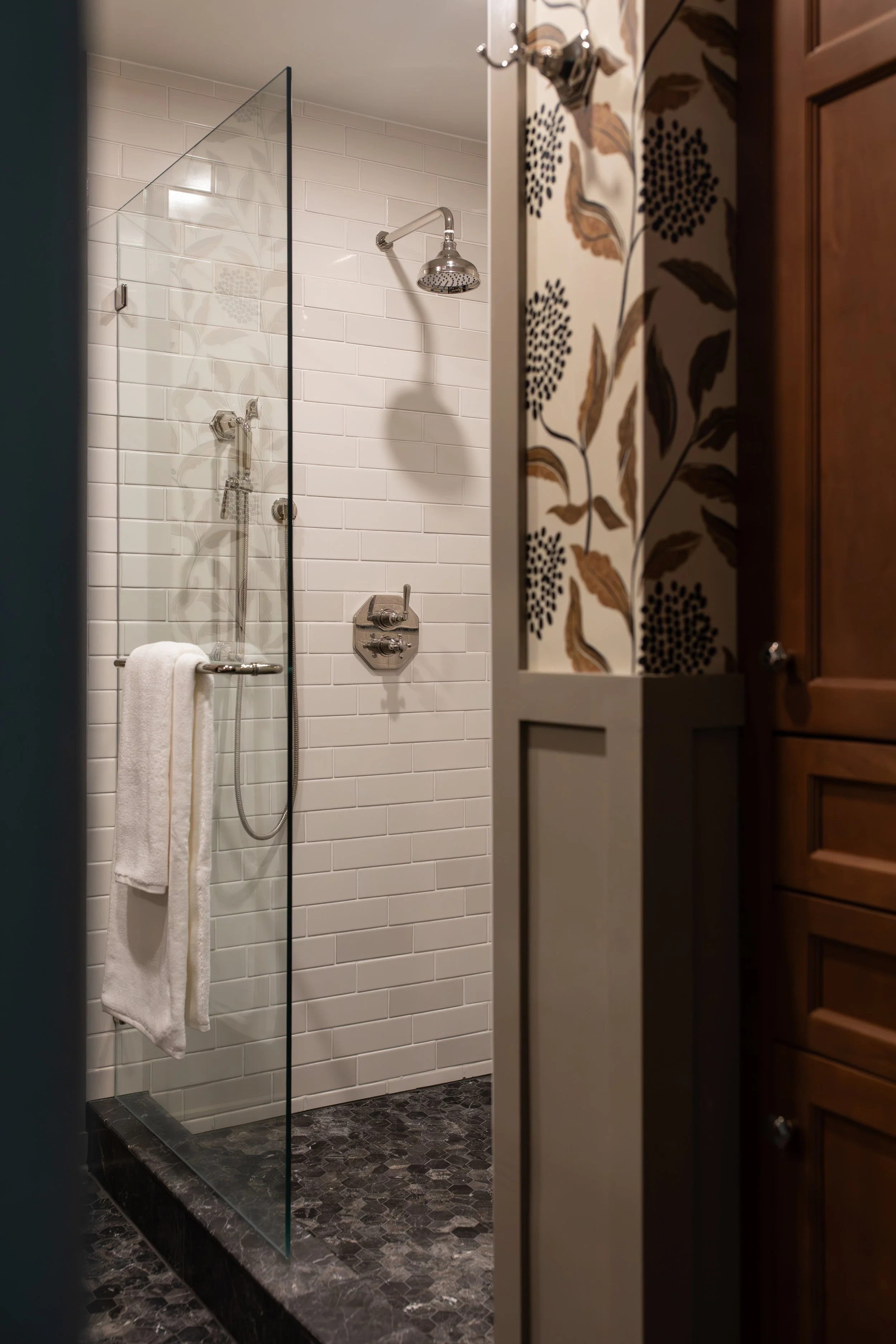 A walk-in shower with white subway tiles, a chrome showerhead, a handheld shower, a glass partition, and a dark pebble-stone floor. A white towel hangs on a rail inside the shower, viewed from an open door with floral and leaf patterned wallpaper and