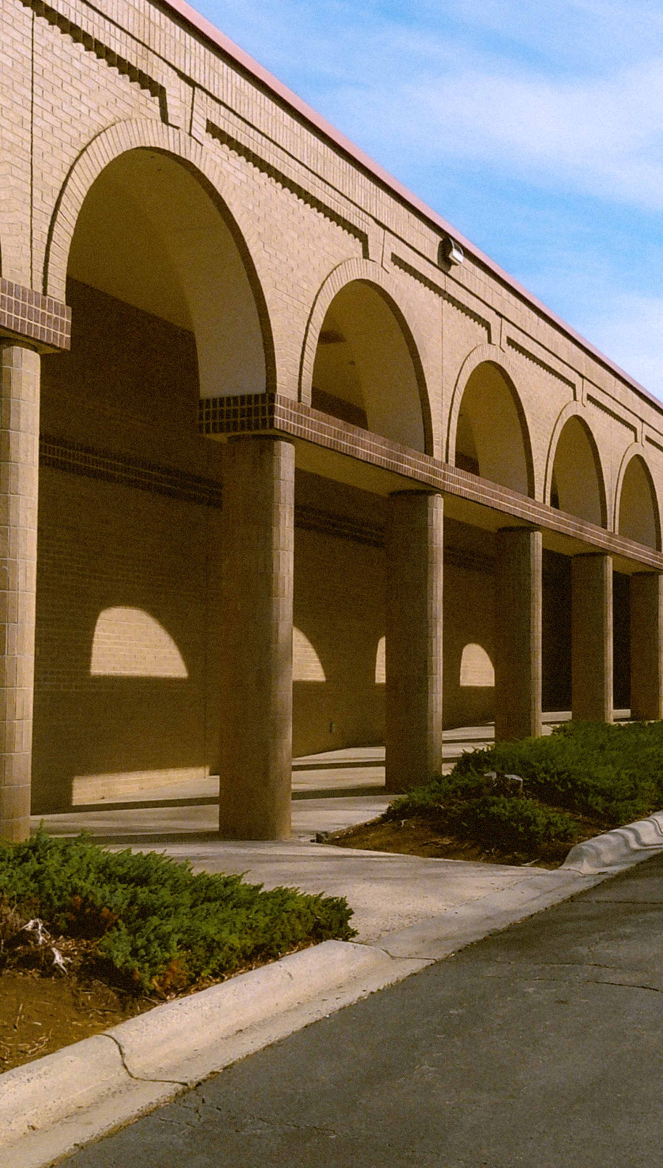 A brick building with arches supported by columns, and a sidewalk with small bushes in front.
