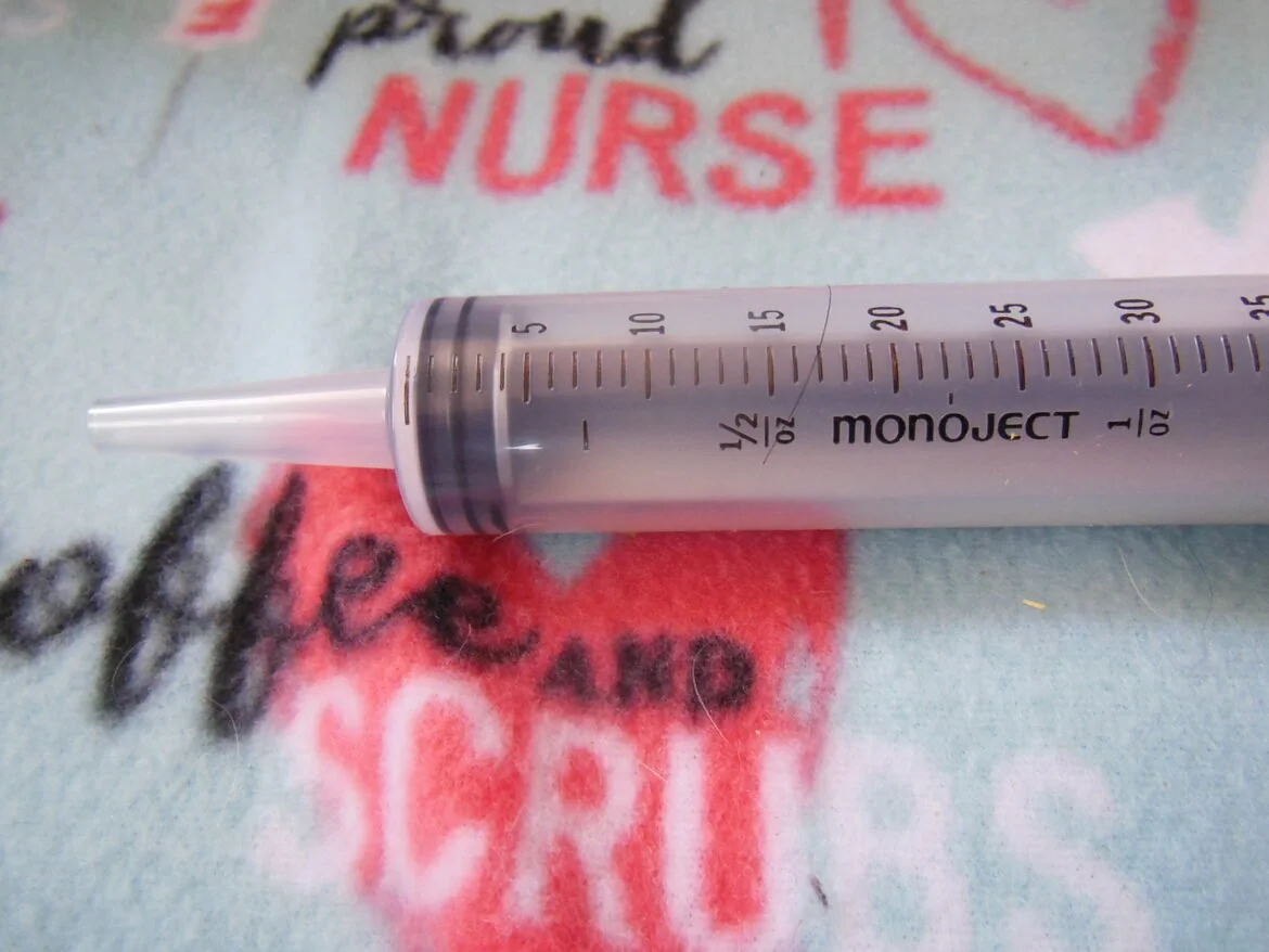 A close-up of a large hypodermic syringe lying on a nurse's uniform with the word 'NURSE' visible in red lettering.