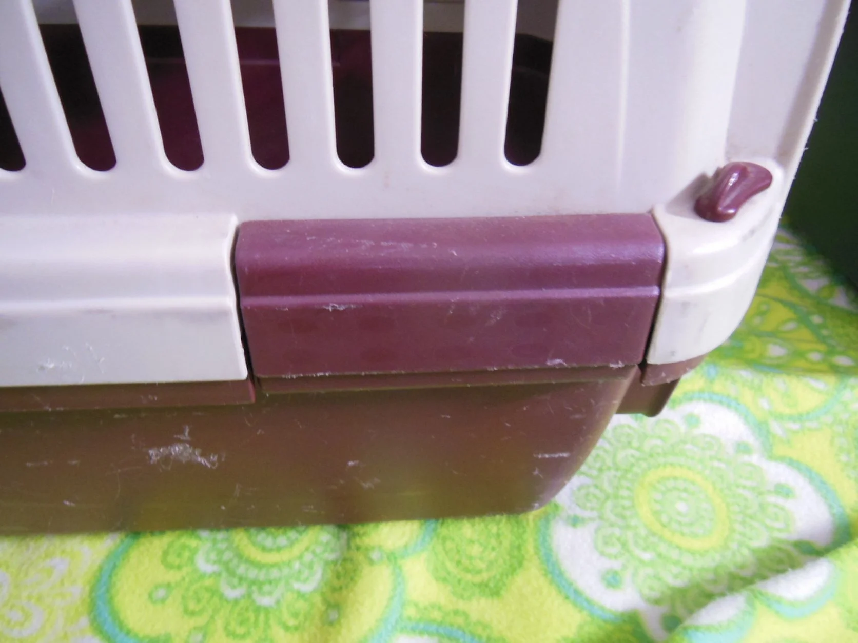 Close-up of a plastic pet carrier with white bars and a purple latch, placed on a colorful patterned fabric.