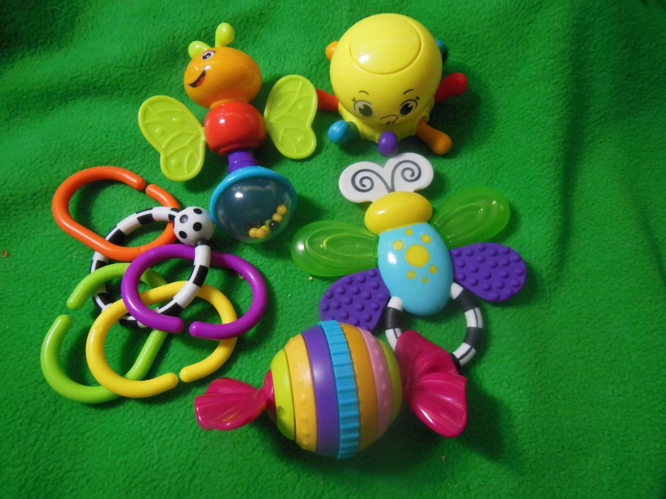 Colorful plastic baby toys on a green surface, including a butterfly, a bee, a butterfly shaped ring toy, and a cluster of interconnected rings.