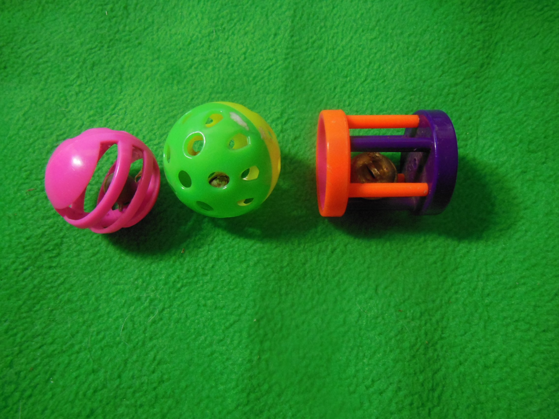 Three colorful cat toys on a green surface: a pink rubber ball with openings, a green perforated plastic ball, and a purple and orange plastic cage with a small bell inside.