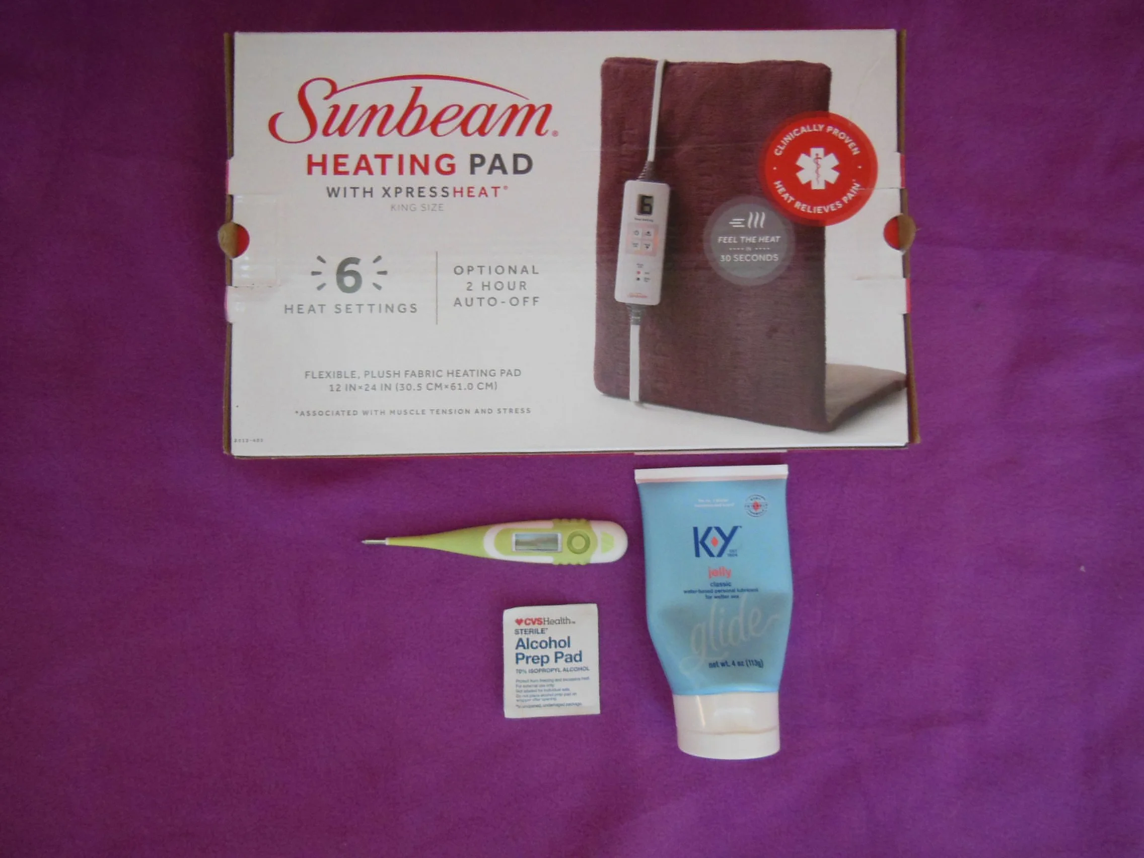 Sunbeam heating pad box, digital thermometer, alcohol prep pad, and tube of KY jelly on purple background.