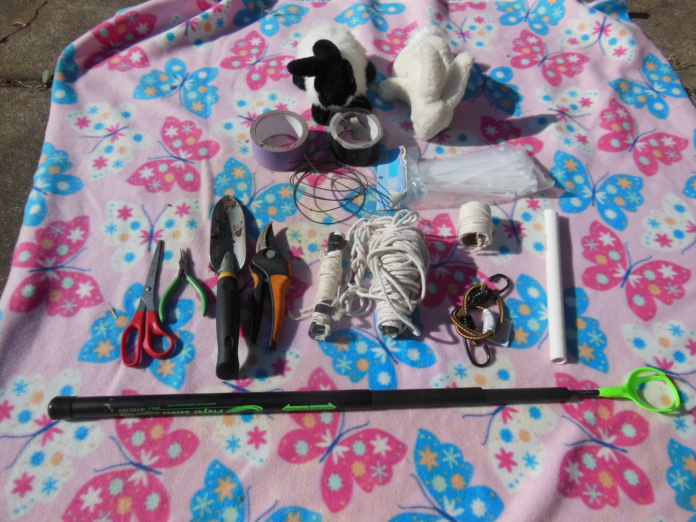 A pink blanket with blue and pink butterfly patterns laid on the ground with various tools and supplies on top. Items include a pair of red-handled scissors, black and orange cutters, a small hand saw, a coil of white rope, rolls of string, a plastic strip, a white plastic stick, a bundle of white feathers in a plastic bag, a black and gray bunny plush toy, a white plush toy, a spool of black thread, a coil of drill wire, and some tape.