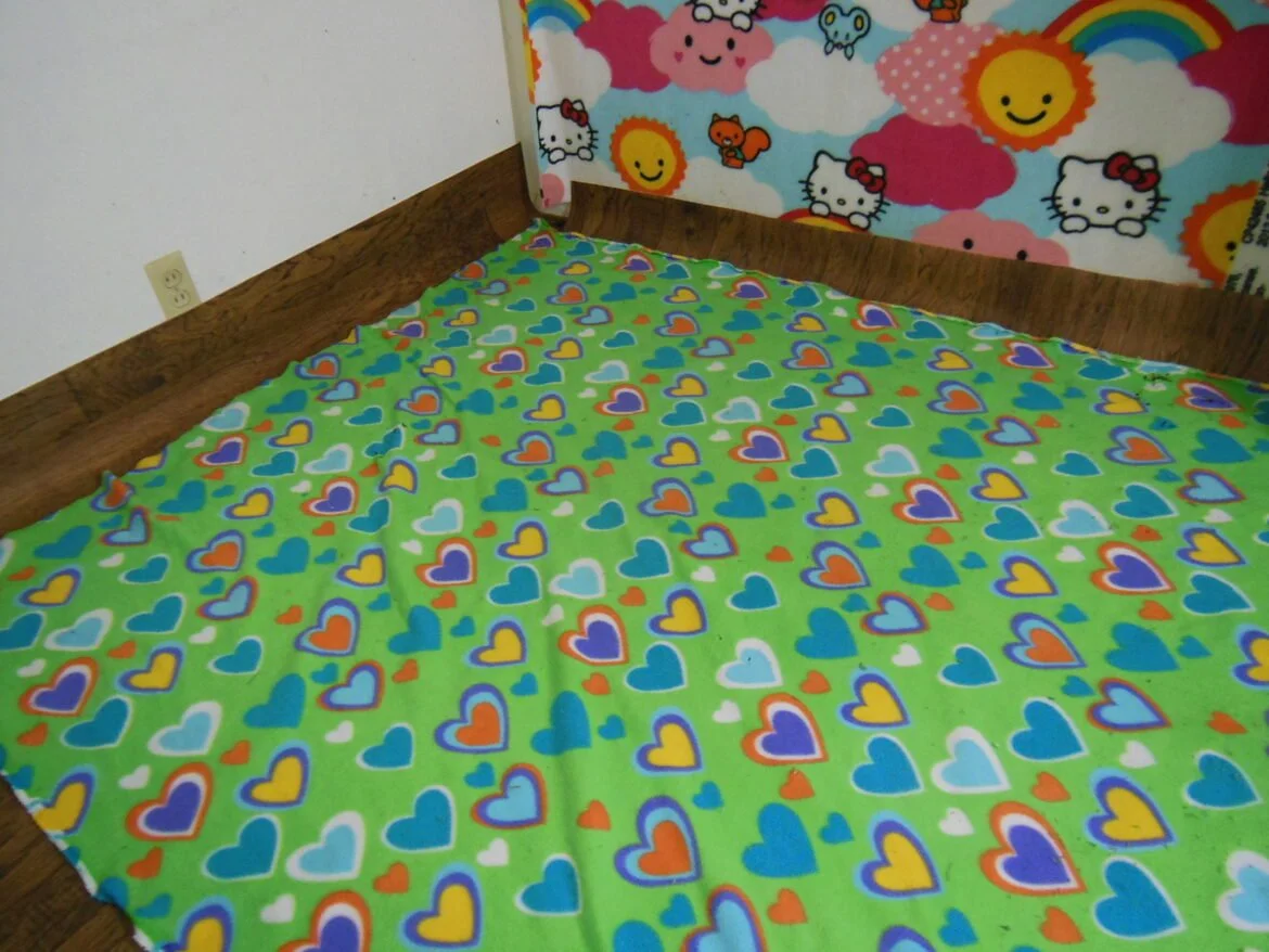 Colorful bedspread with blue, yellow, orange, white, and purple hearts on a green background, against a wall with Hello Kitty and rainbow-themed wallpaper.