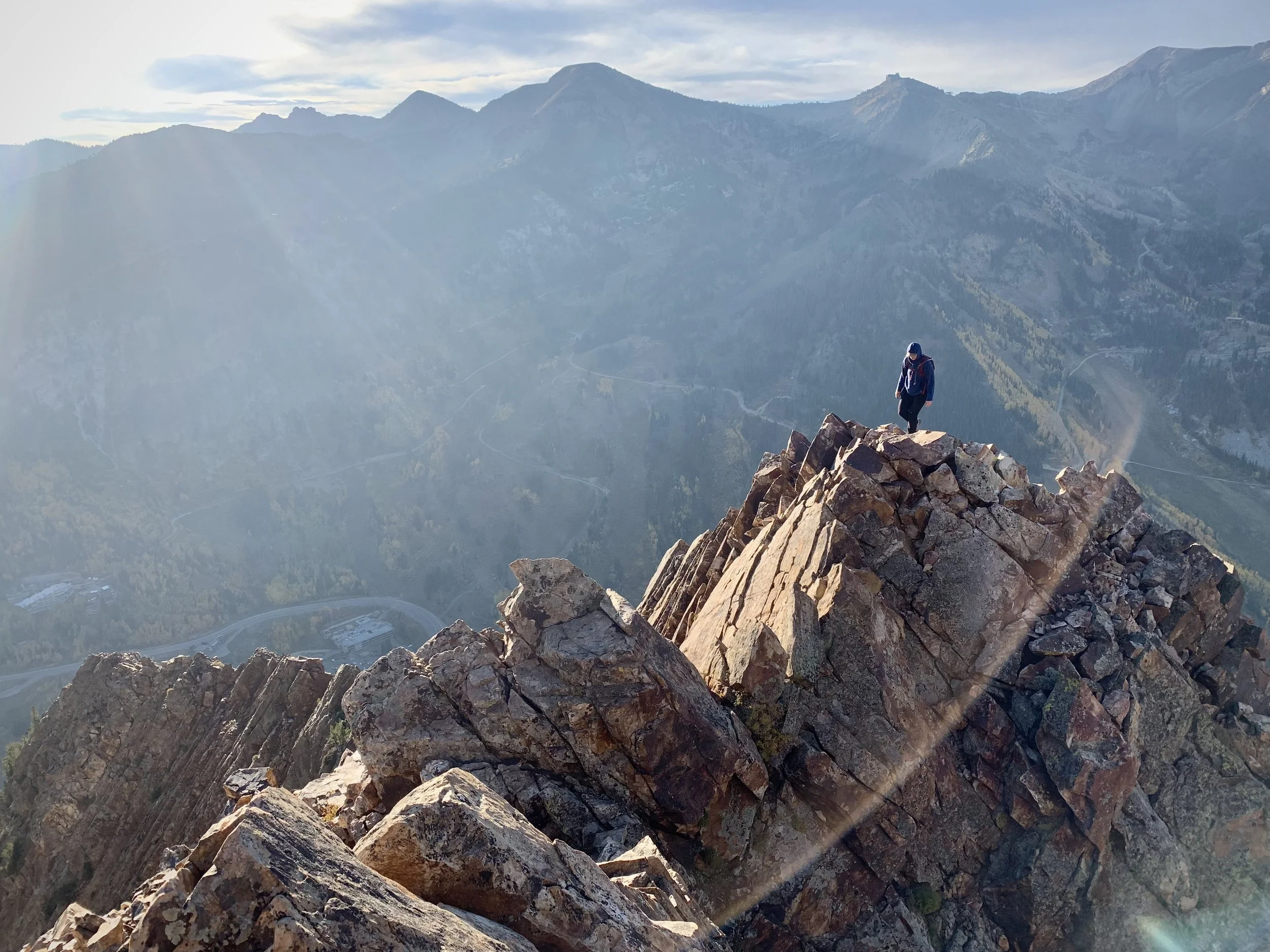 A person wearing a blue jacket and backpack standing on rocky mountain peak with a view of mountain ranges and a valley below.