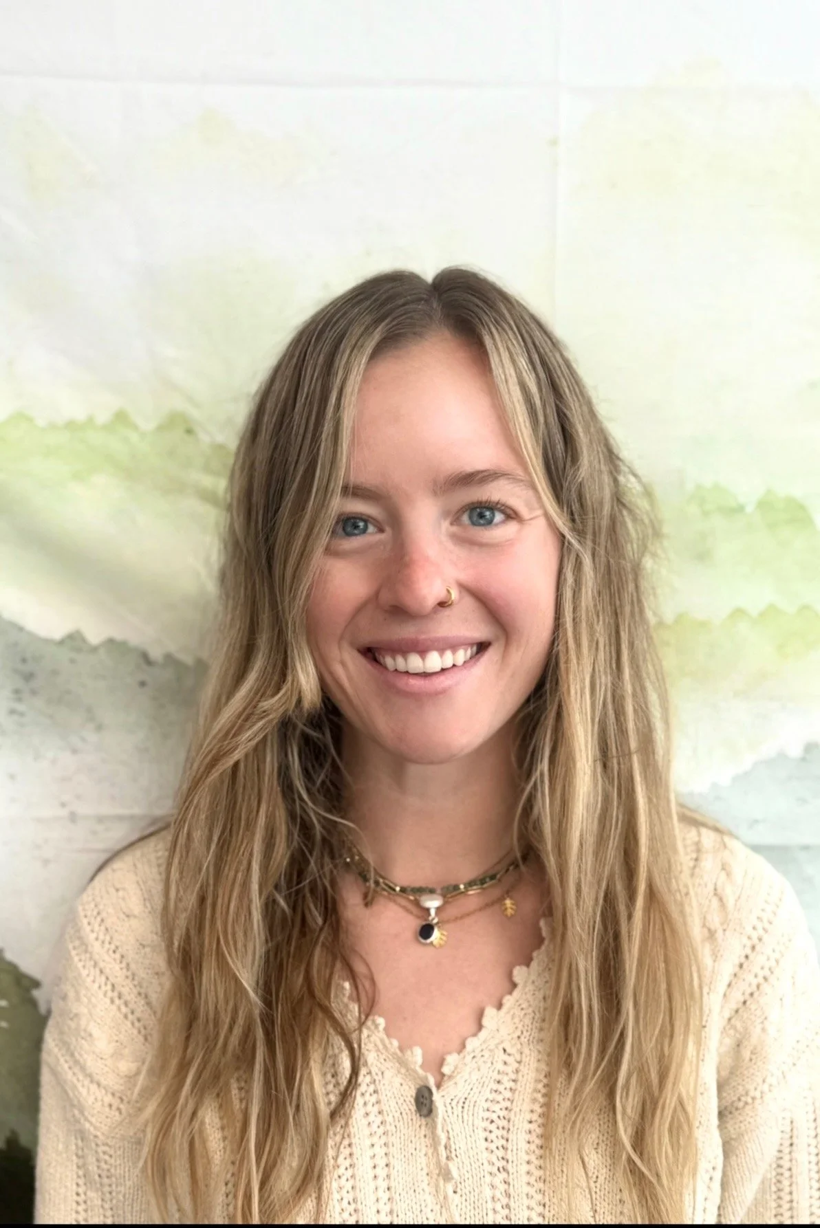 A young woman with long wavy blonde hair, blue eyes, and a nose ring, smiling at the camera, against a watercolor background.