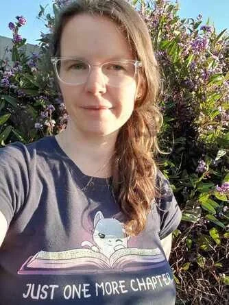 A woman with glasses taking a selfie outdoors in front of purple flowering bushes on a bright day.