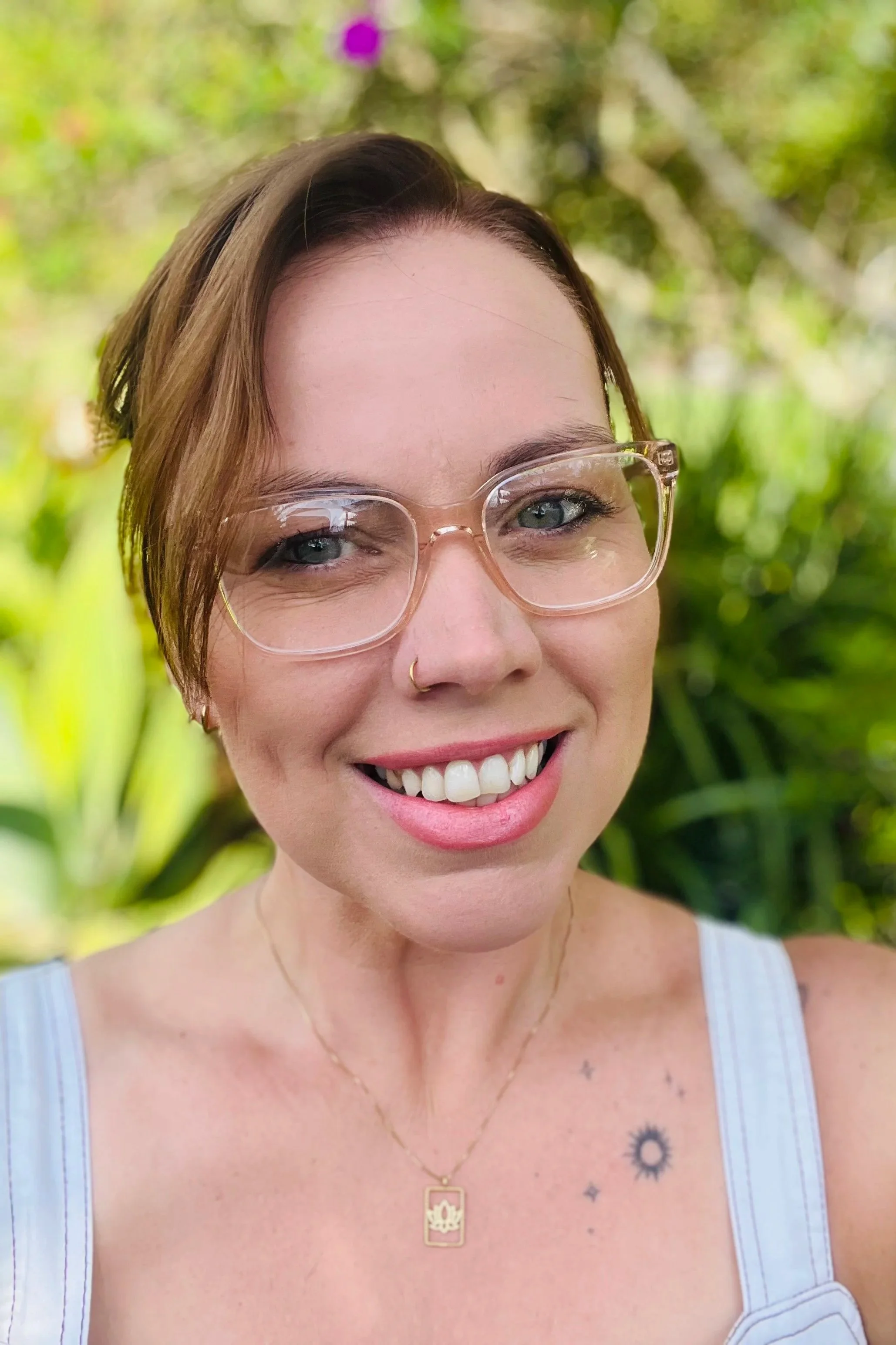 Close-up of a smiling woman with glasses, short brown hair, a nose ring, tattoos on her shoulder, wearing a tank top and a gold necklace, outdoors with green foliage in the background.