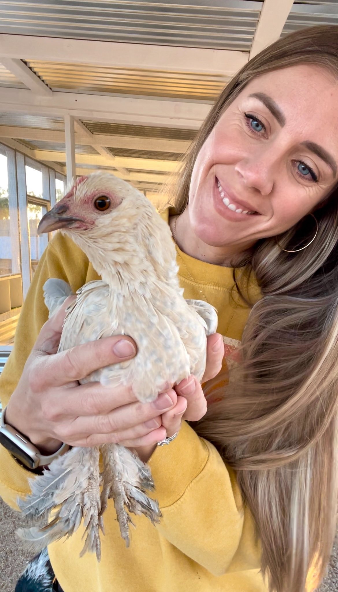 A woman smiling and holding a young, light-colored chicken inside a building with a metallic ceiling and windows.