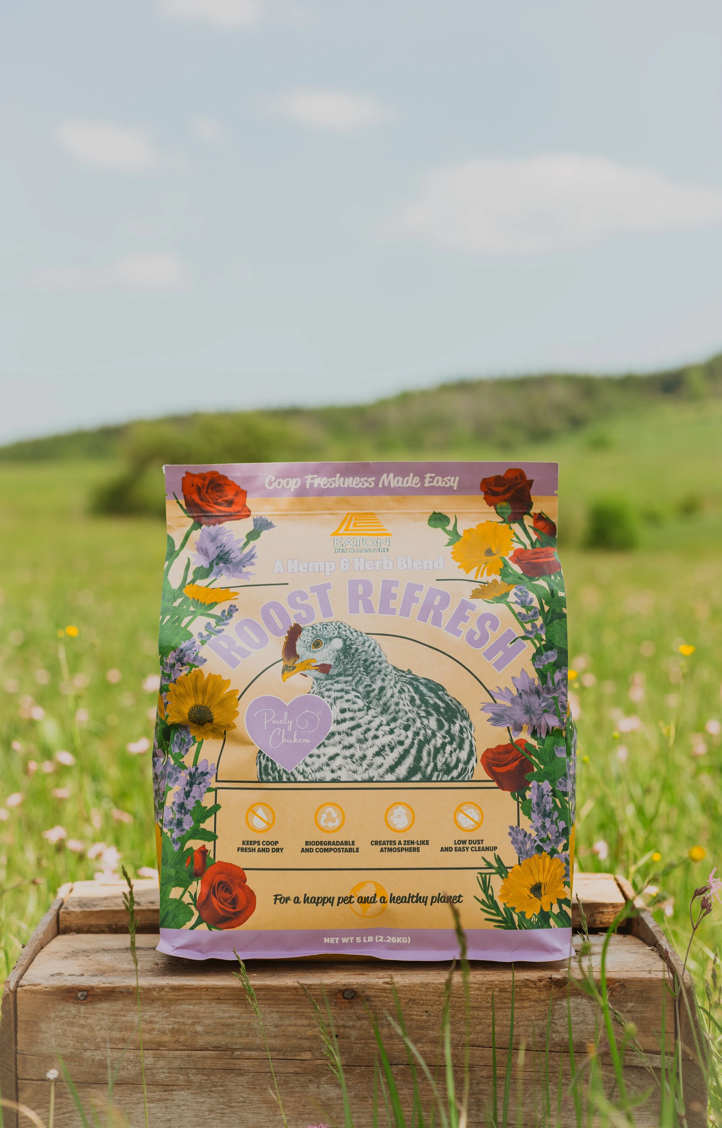 A bag of Chicken Feed labeled 'Roost Refresh' standing on a wooden surface in a grassy field with hills and a blue sky with clouds in the background.