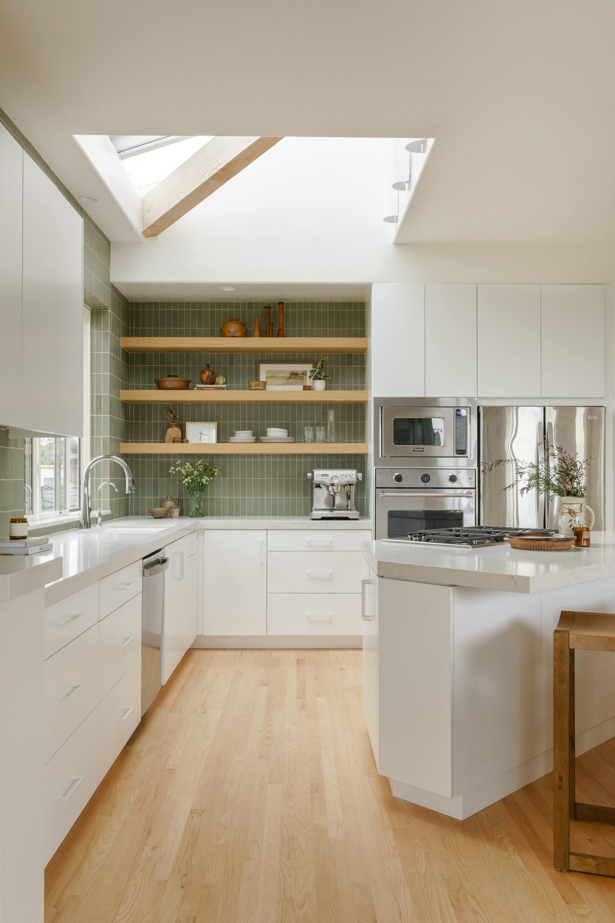 Modern kitchen with white cabinets, wooden open shelves, green tile backsplash, stainless steel appliances, and natural light from a skylight.