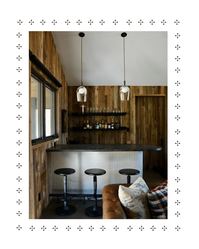 Interior view of a small bar area with a wood-paneled wall, a window on the left, a black countertop with three black bar stools, a wall shelf with bottles and glasses, and a beige and patterned pillow on a brown leather couch in the foreground.