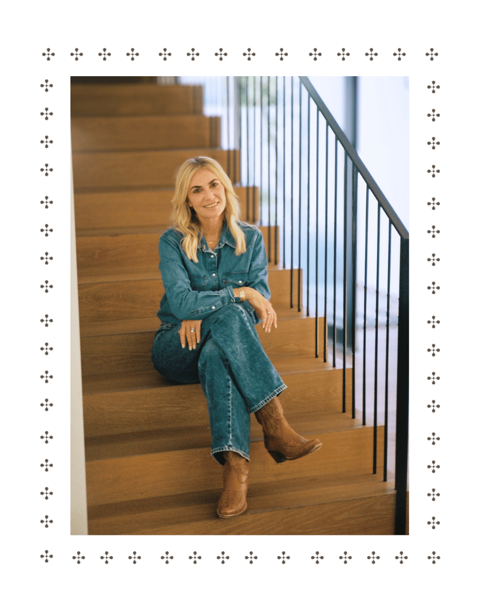A woman with blonde hair sitting casually on wooden stairs, wearing a denim jacket, jeans, and cowboy boots, smiling at the camera.