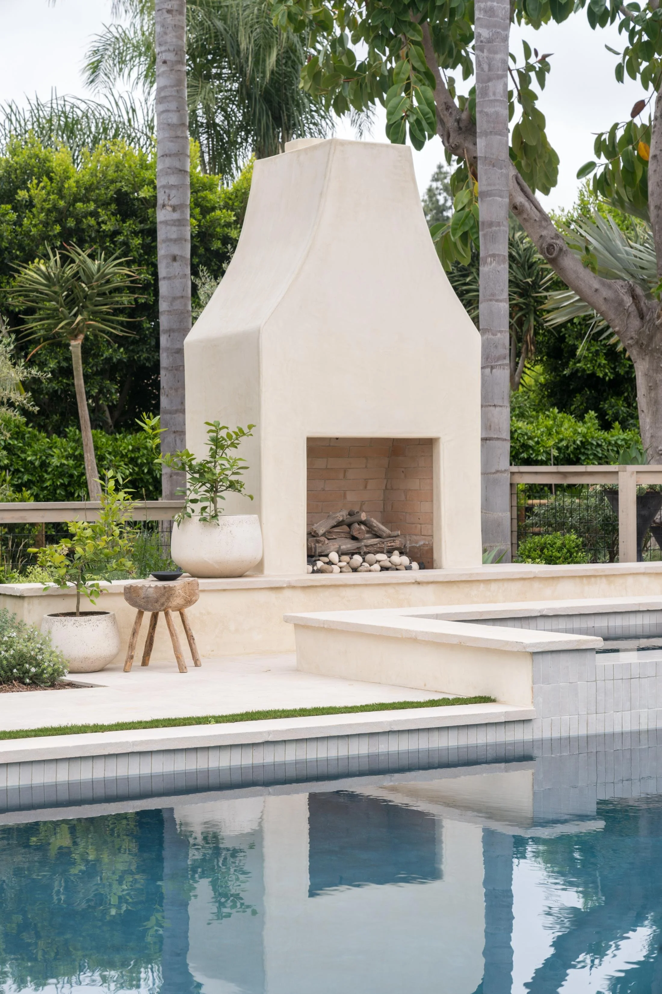 Outdoor fireplace with a pool in foreground, surrounded by lush green plants and trees.