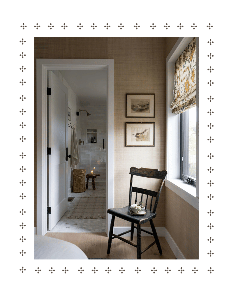 A cozy bedroom corner with a wooden rocking chair holding a small book and a bowl, near a window with floral curtains and framed bird illustrations, leading to a bathroom with a candle on a small table and wicker basket.