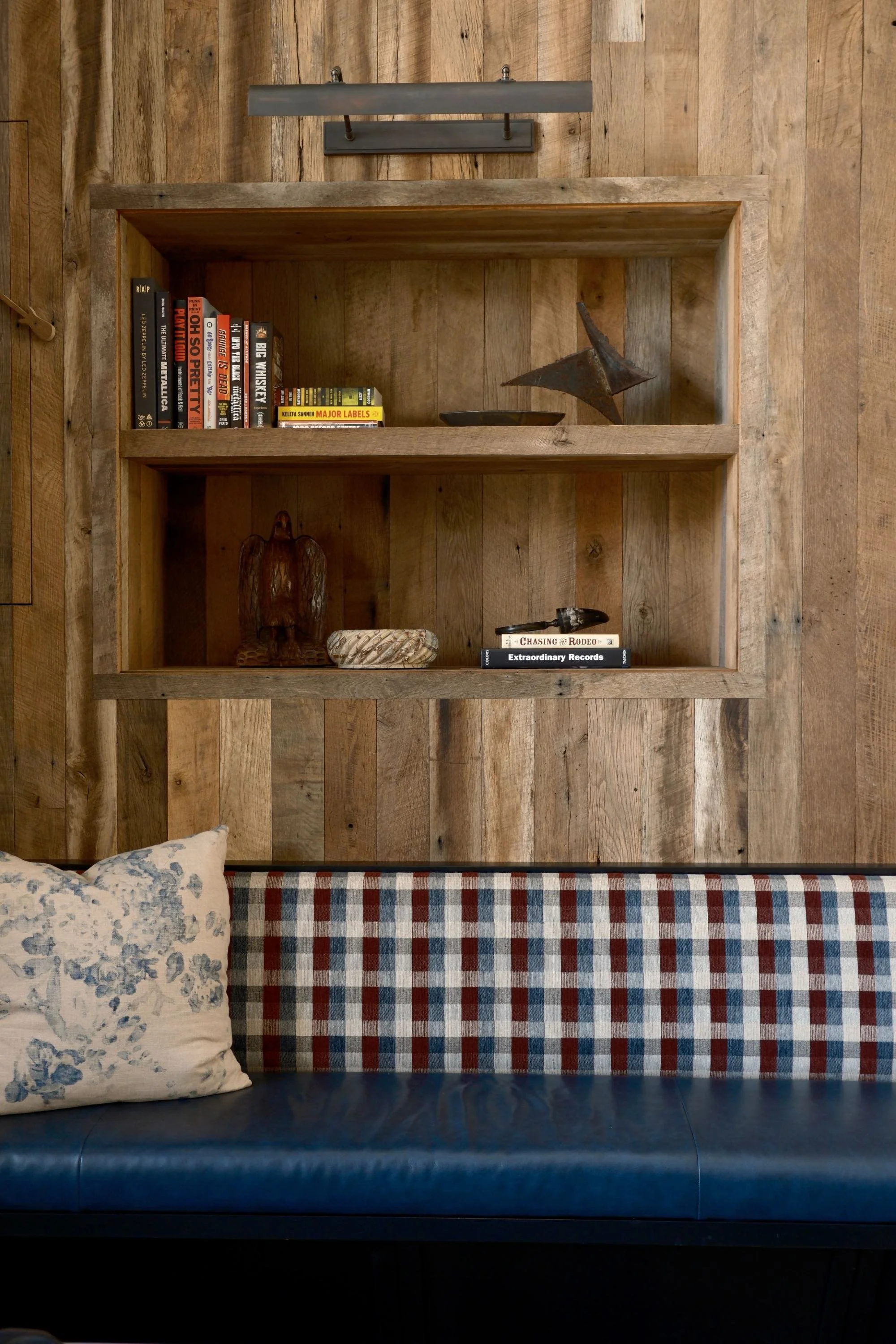 Wooden wall with built-in shelves holding books and decorative objects, part of a room with a blue bench, plaid and floral pillows.
