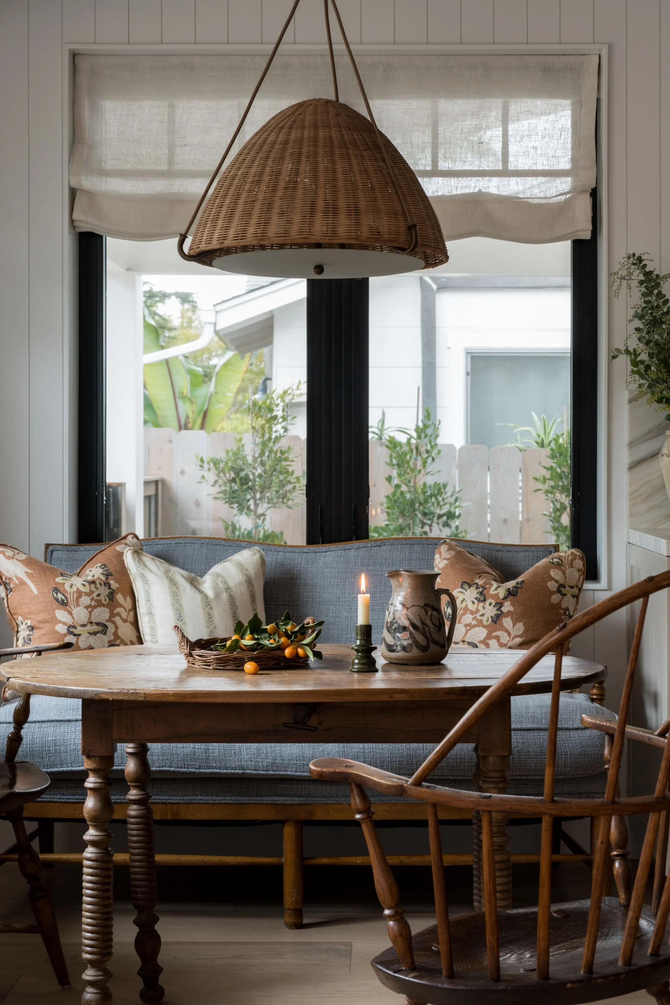 Cozy dining nook with a wooden table, vintage rocking chair, and a woven hanging light fixture. Items on the table include a lit candle, a decorative pitcher, and a basket of small oranges. The background shows large windows with a view of a backyard garden.