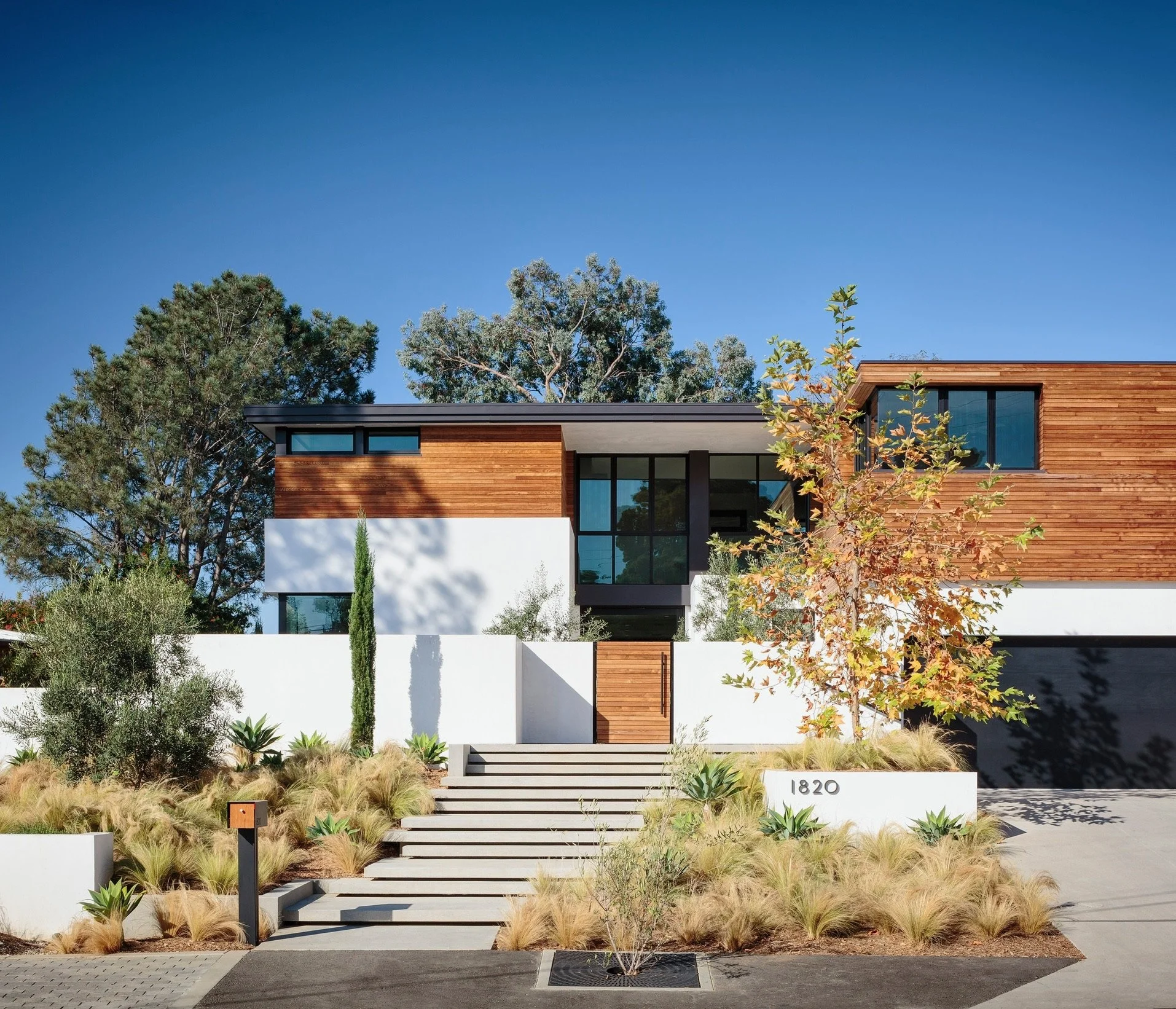 Modern house with wooden and white exterior, glass windows, landscaped front yard with trees and plants, concrete stairs, and house number 1820.