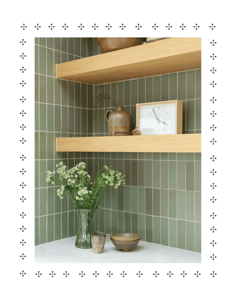 Green tiled kitchen corner with two wooden shelves, a vase of white flowers, and decorative pottery on countertop.