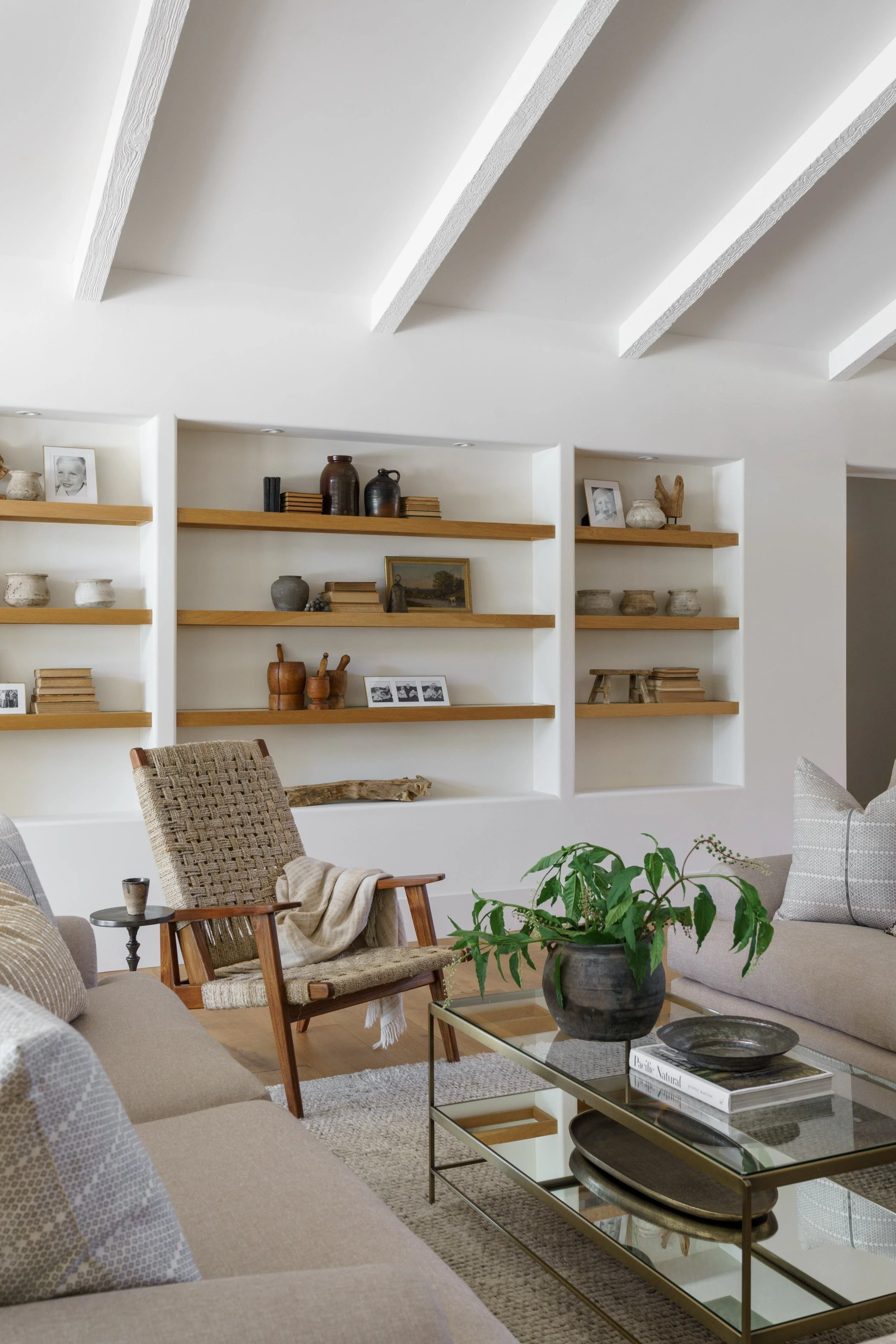 Living room with beige sofas, a woven armchair, a glass coffee table with books and decorative bowls, a plant, and built-in shelves with vases, books, and framed photos.