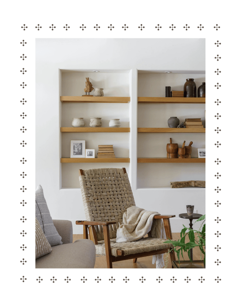 Living room with built-in white shelves holding pottery and decor, a woven armchair with a blanket, part of a gray sofa with pillows, and a potted plant.
