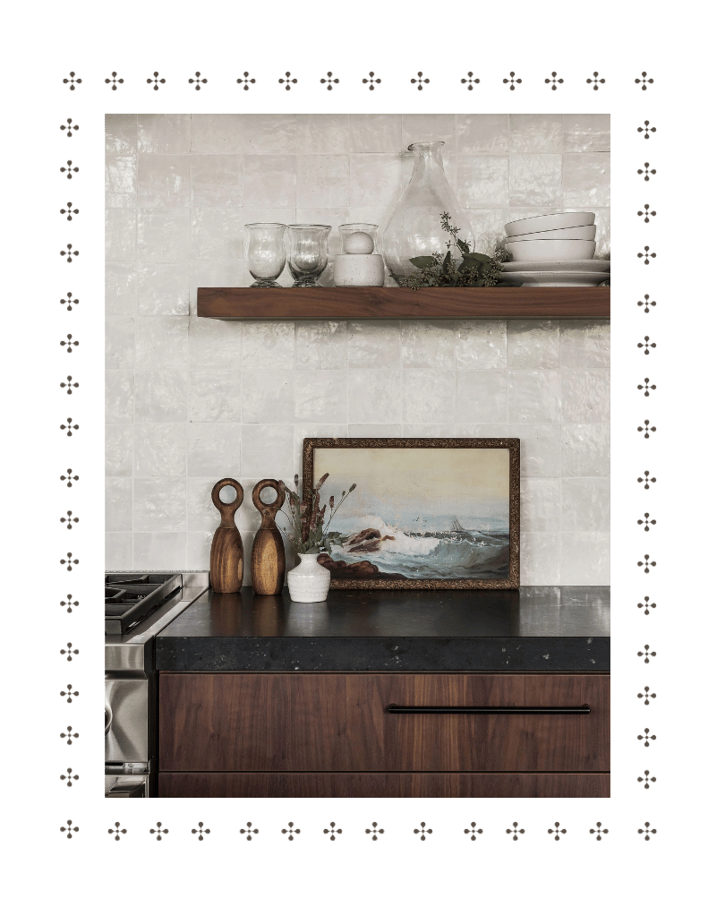 Kitchen countertop with a painting, vases, and decorative items, and a wooden shelf with glassware and dishes above, against a tiled wall.