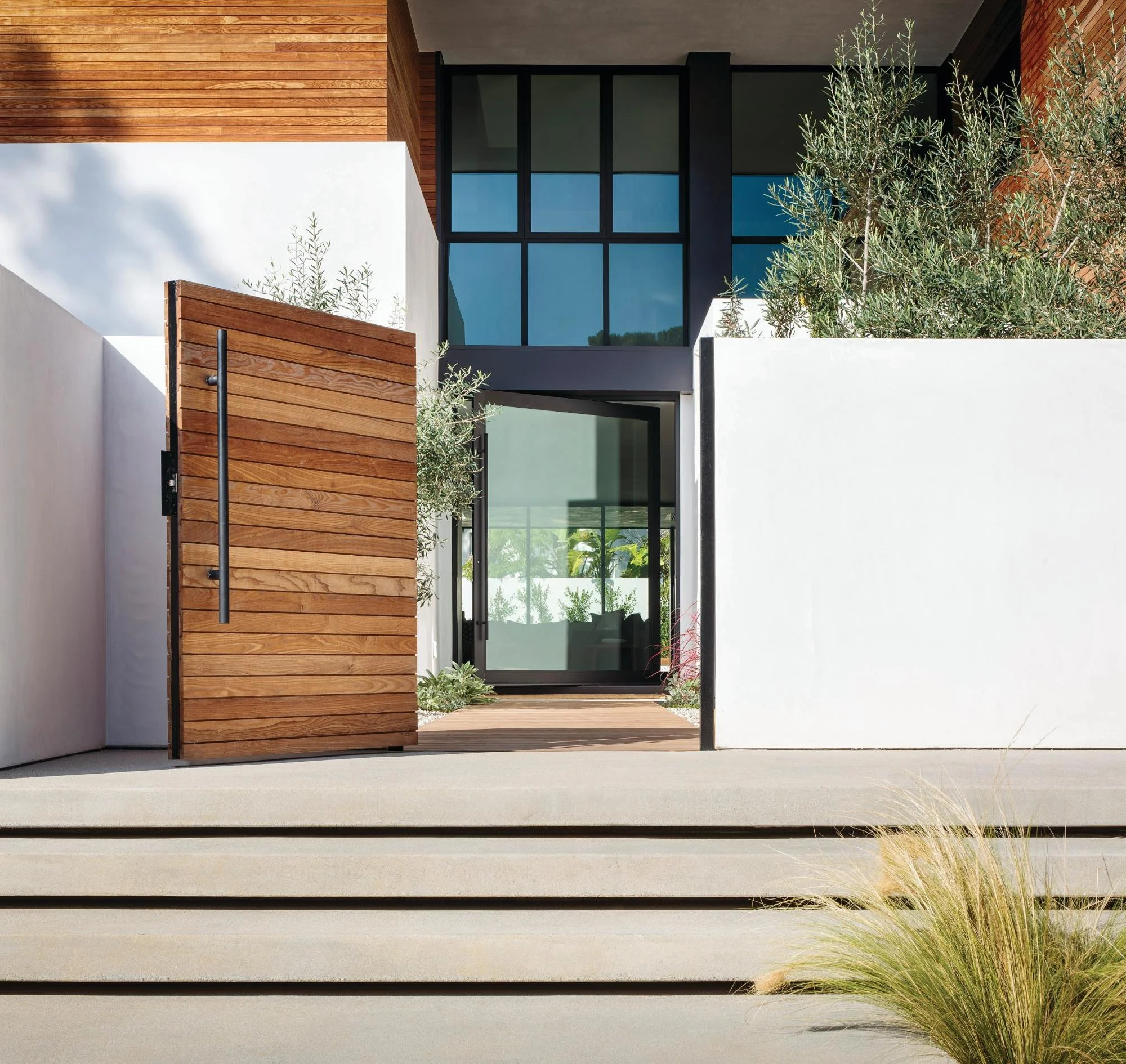 Modern house with wooden and black-framed glass exterior, white walls, and a large open wooden gate, surrounded by greenery.