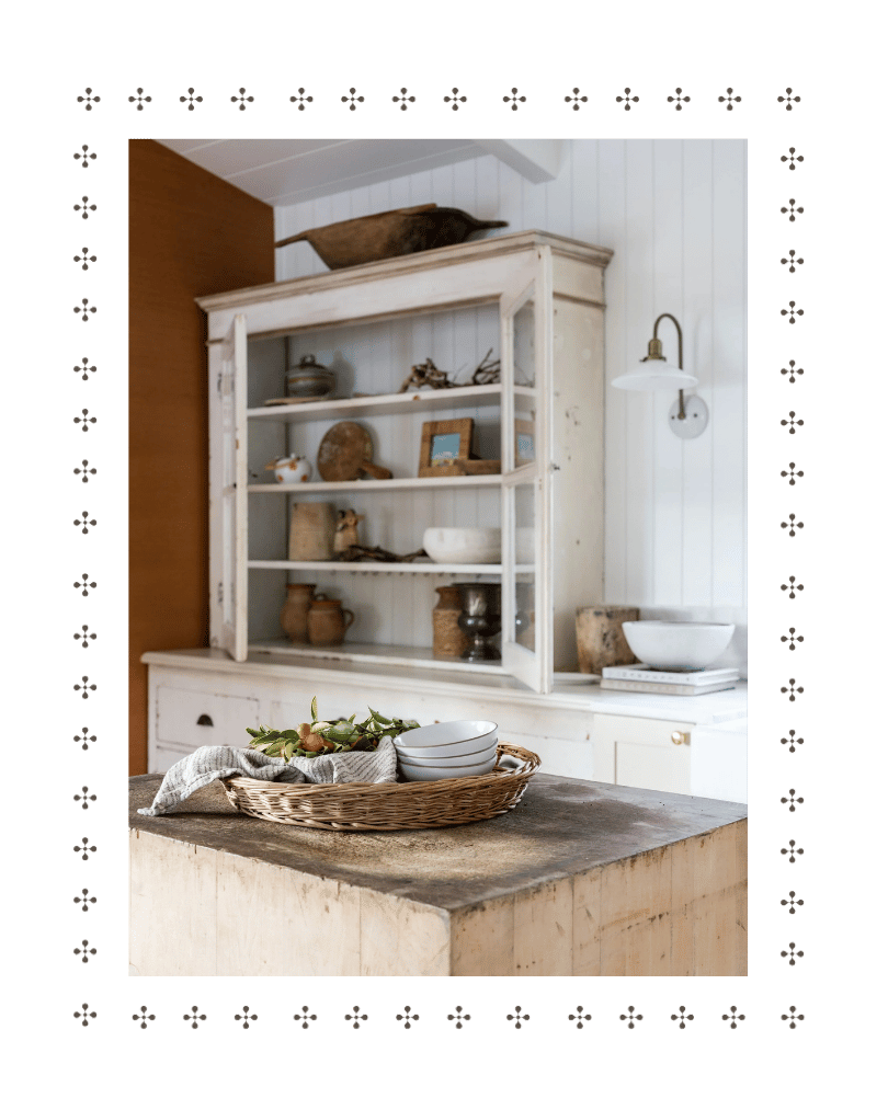 A cozy rustic kitchen with a light-colored open shelving unit displaying various pottery and decorative items. A wooden island with a wicker basket containing white bowls and greenery in the foreground. A white wall-mounted lamp and a small white bowl on stacked books are on the right side.