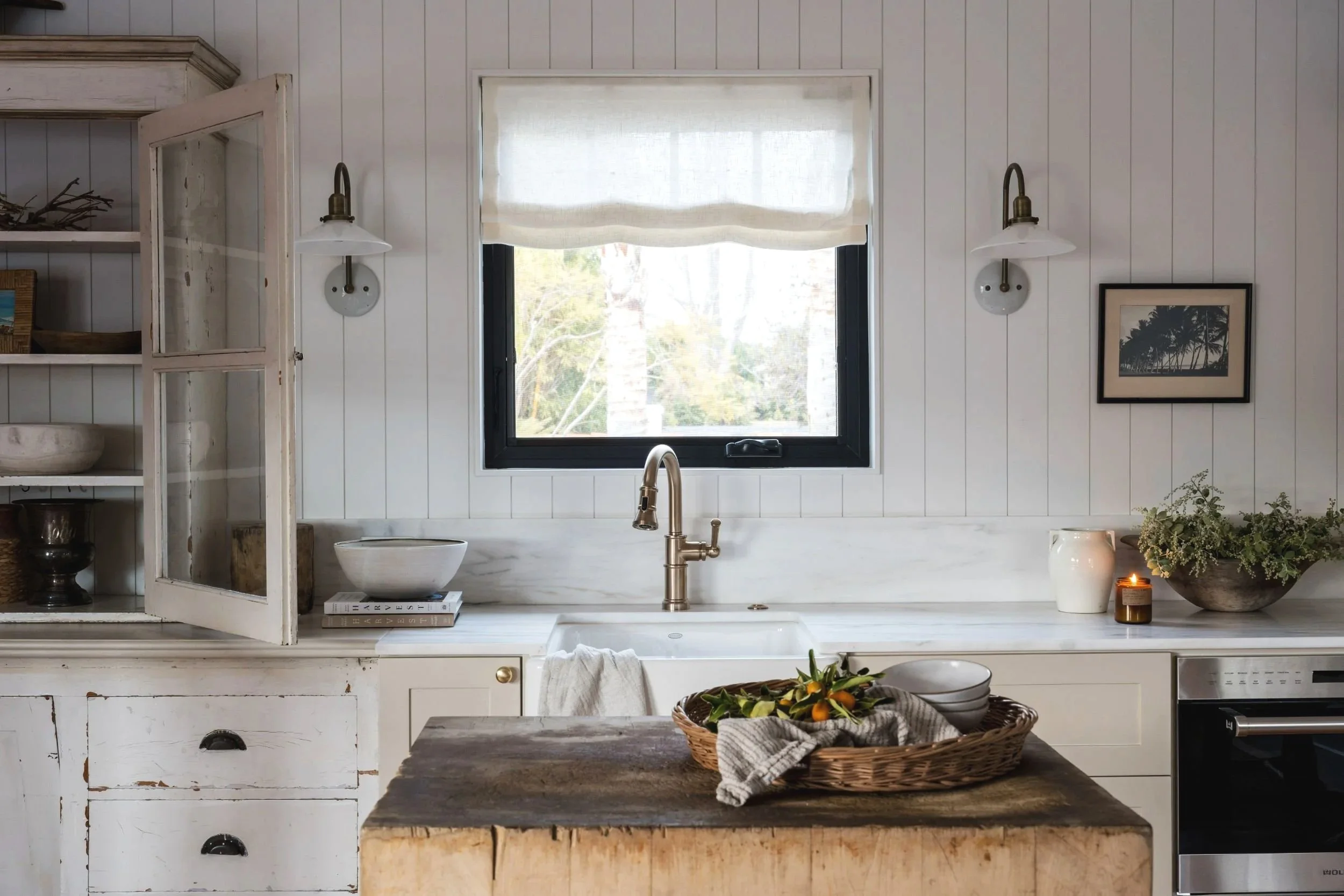 Cozy kitchen with white wooden panel walls, a window with a roman shade, built-in shelves, a black-framed window, a farmhouse sink with a brushed bronze faucet, a wooden island with a basket of fruit, decorative bowls, a candle, a potted plant, and wall-mounted sconces.