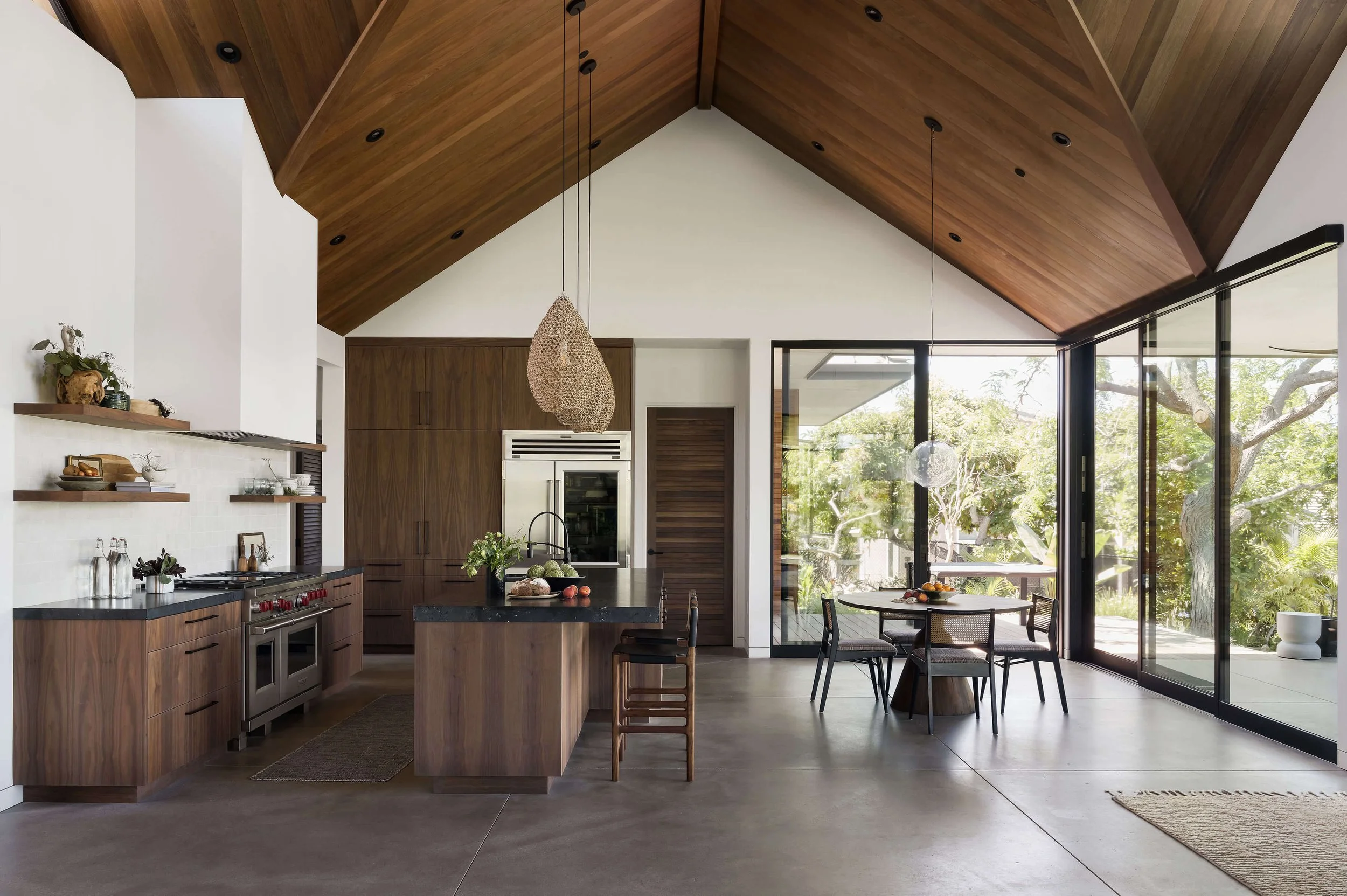 Modern kitchen with wooden cabinets and high vaulted ceiling, open to a dining area with large glass sliding doors revealing trees outside.