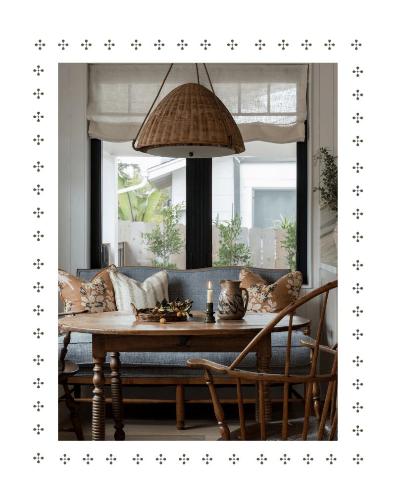 A cozy dining room with a wooden table, a candle, and ceramic vases, surrounded by wooden chairs, in front of large window with plants outside.