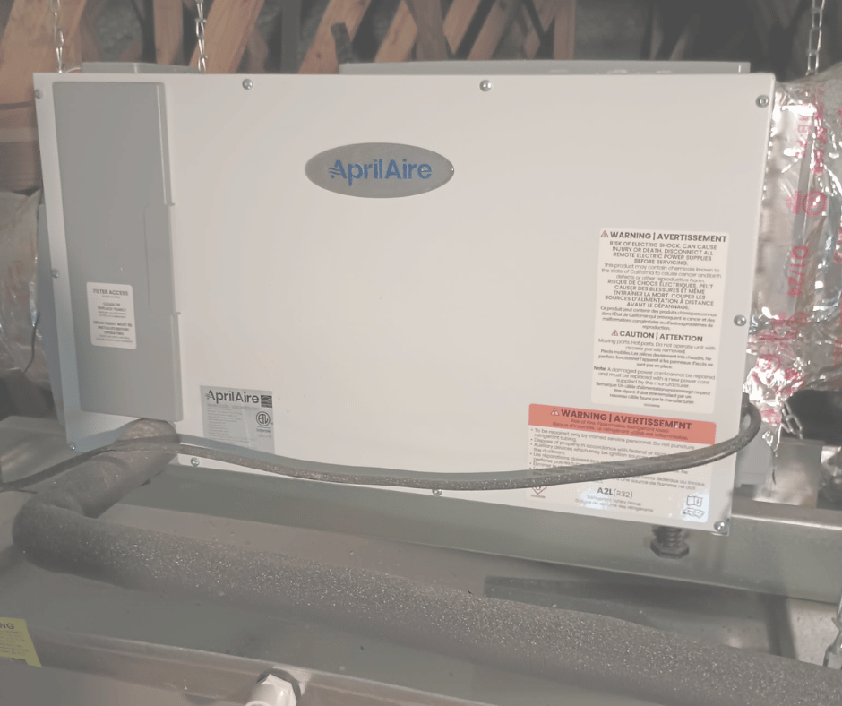 A white rectangular HVAC unit labeled AprilAire, installed in an attic space with wooden beams, with warning labels and a flexible pipe attached.