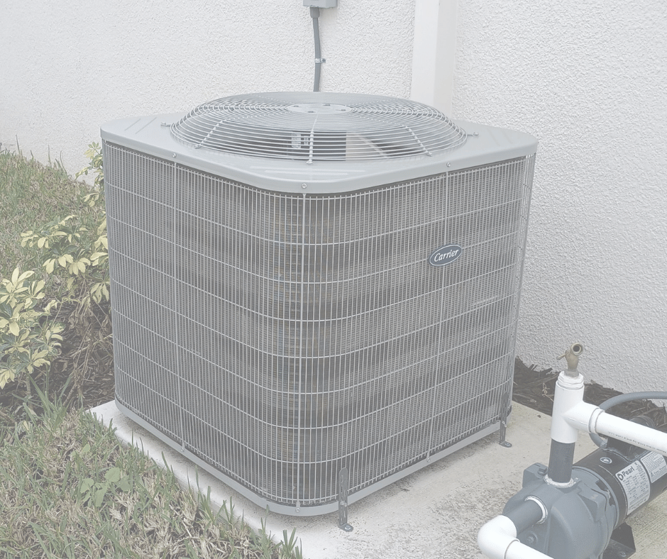 A gray Carrier air conditioning unit outside a building on a concrete slab, next to a garden and a white wall.