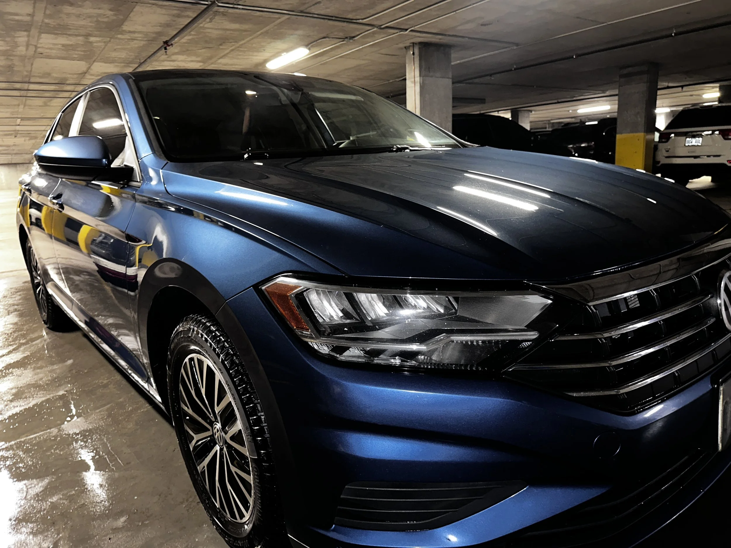 A blue Volkswagen sedan parked in an indoor parking garage with concrete ceiling and other vehicles in the background.