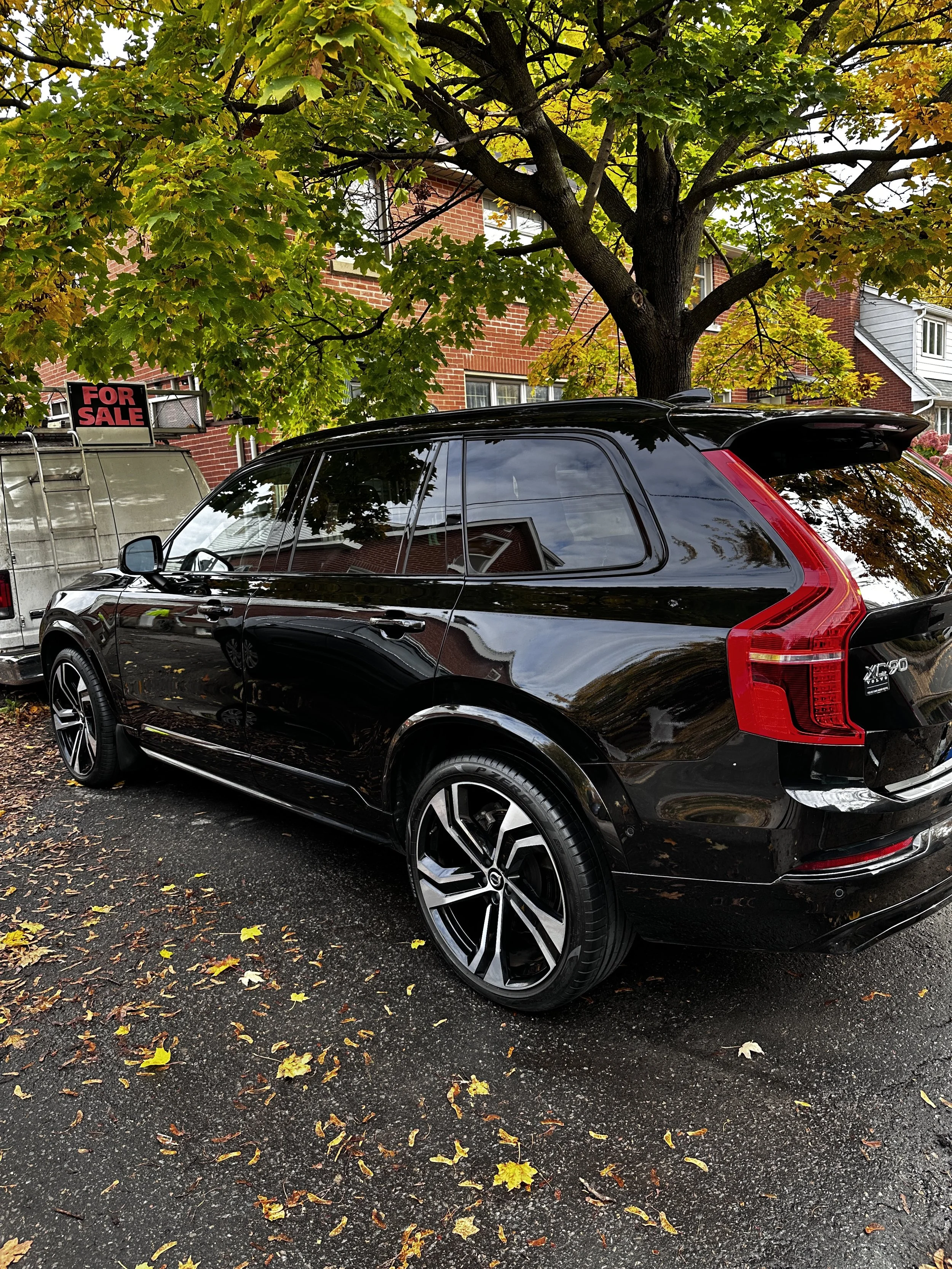 A black SUV parked on a street with fallen leaves, under a large tree with green and yellow leaves, a house with brick walls in the background, and a 'For Sale' sign on a trailer nearby.