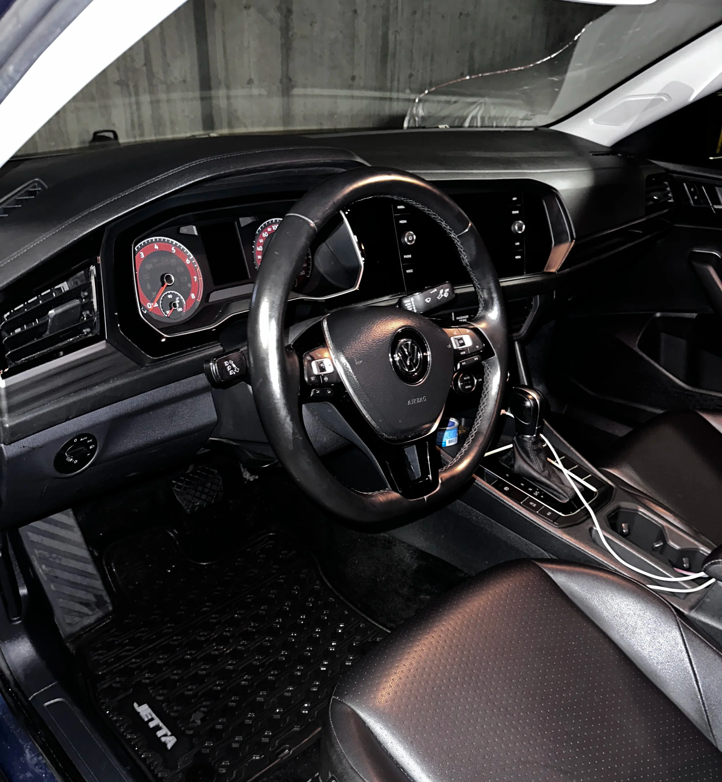 Interior of a Volkswagen vehicle, showing the steering wheel, dashboard with speedometer and tachometer, gear shift, and a charging cable connected to a port near the center console.