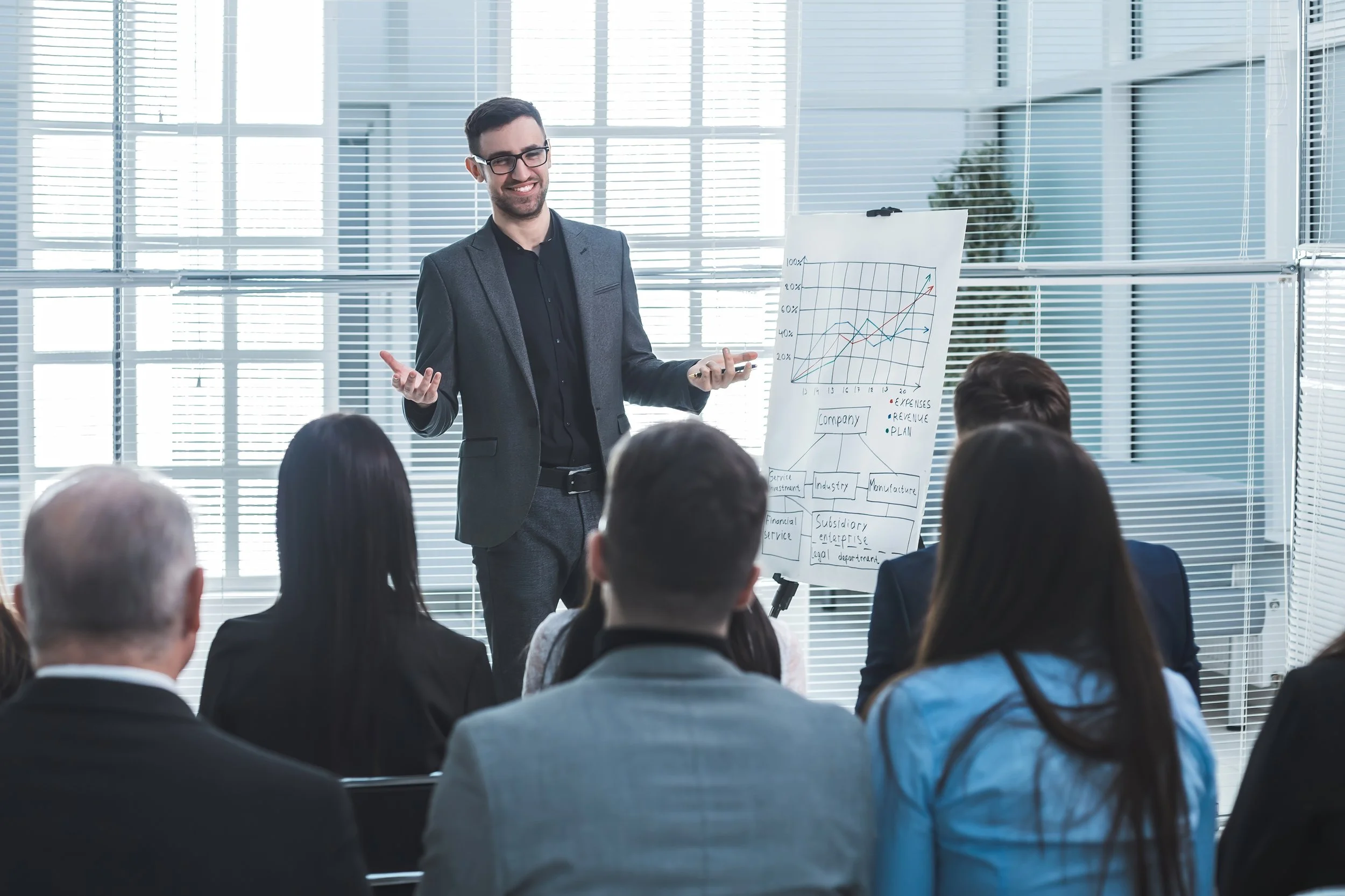 A man in a suit giving a presentation in front of an audience, with a chart on a whiteboard showing increasing lines and notes about revenue and expenses.