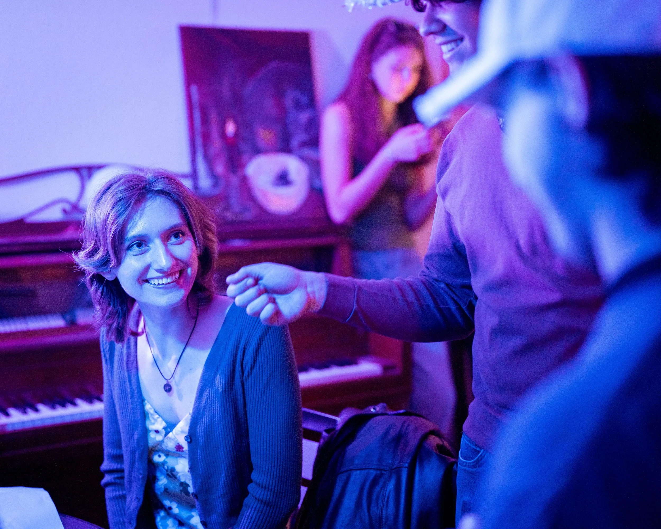 People socializing at a piano bar with colorful lighting, focusing on a woman with short red hair smiling at a man, with women in the background using their phones.