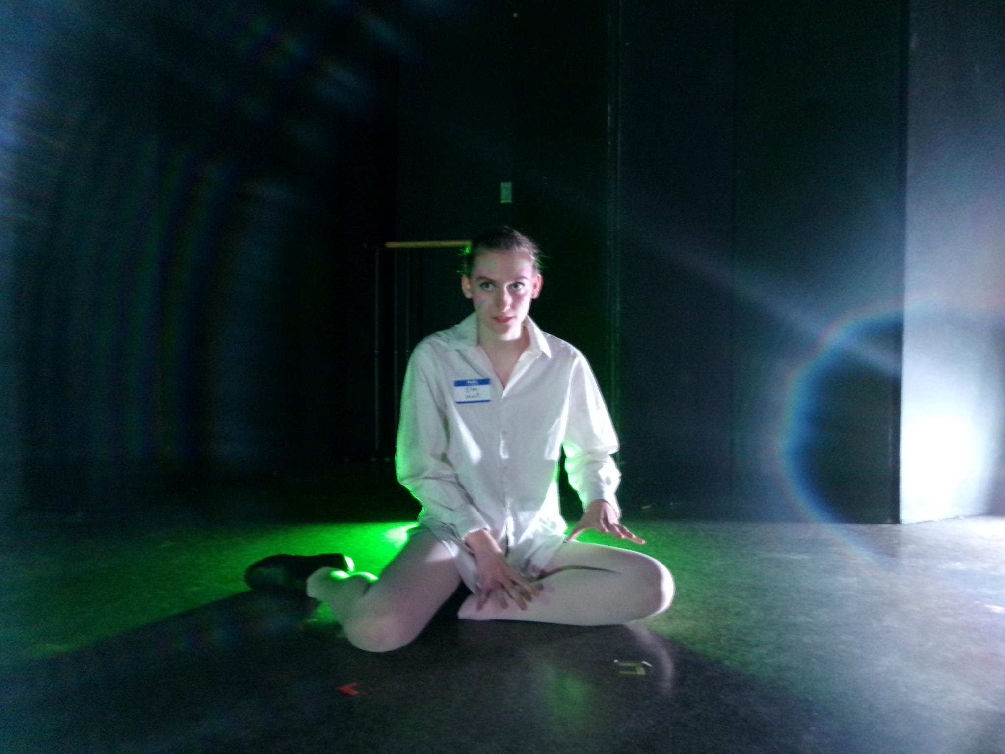 A woman in a dance or theatrical costume kneels on the floor in a dark room with dramatic lighting, including green and white light effects.