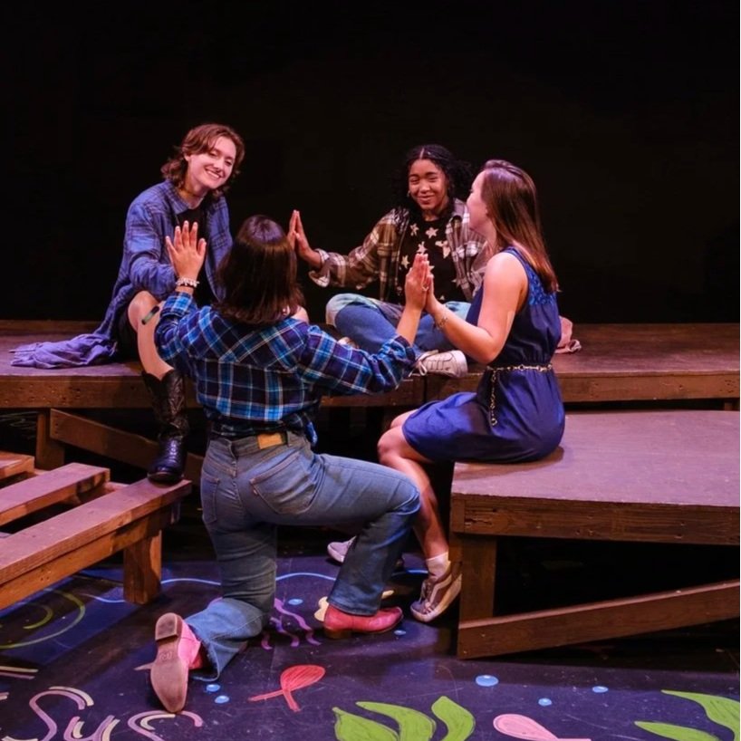 Four women are playing on a small stage with a dark background. They are sitting and kneeling, facing each other, and giving high fives. One woman is smiling, and they all seem to be enjoying a theatrical performance or rehearsal.
