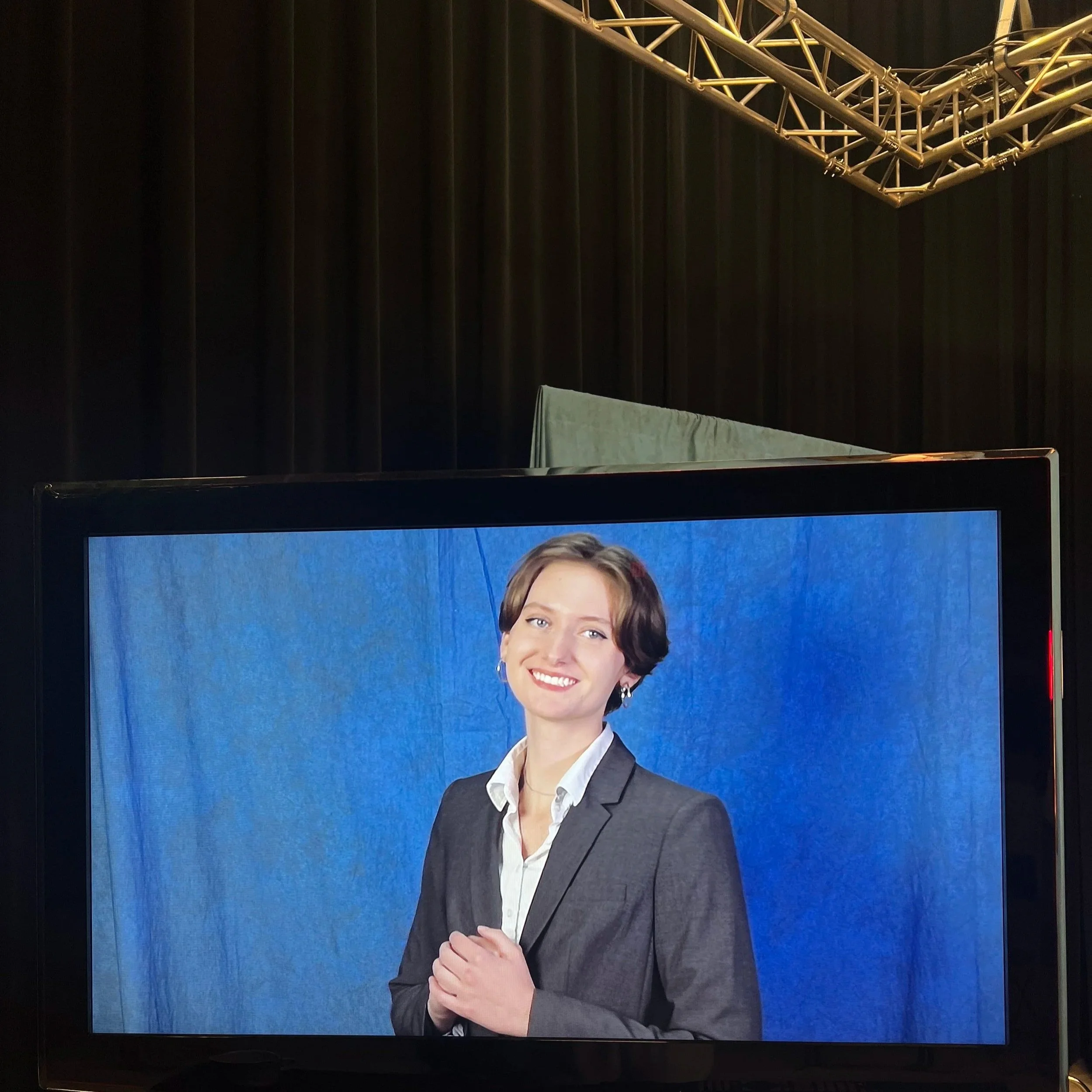 A woman with short brown hair wearing a gray blazer and white shirt, smiling, with hands clasped in front of her, shown on a monitor against a blue background.