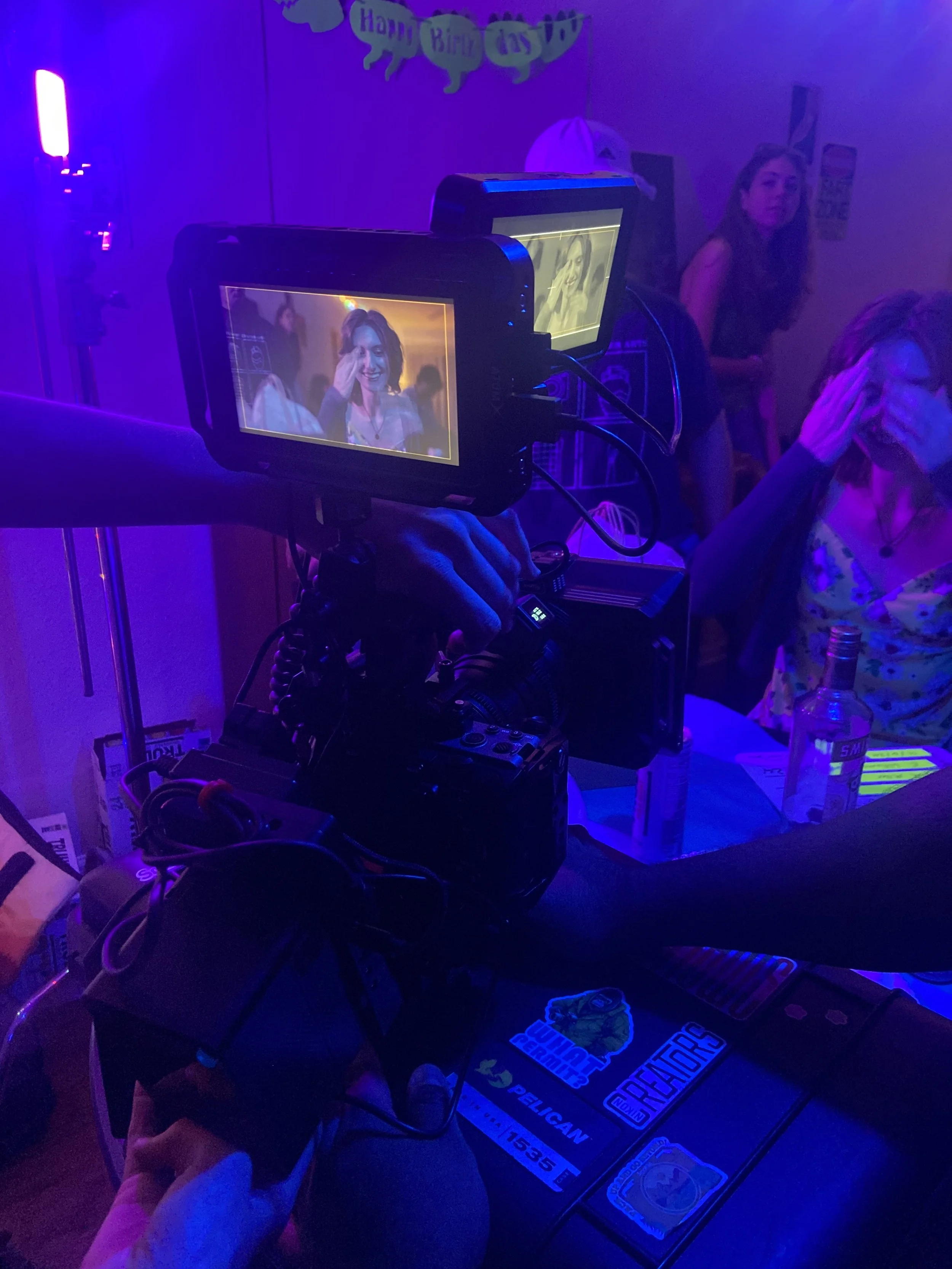 A film camera on a tripod filming a woman smiling at a party in a room decorated with a "Happy Birthday" banner and purple lighting.