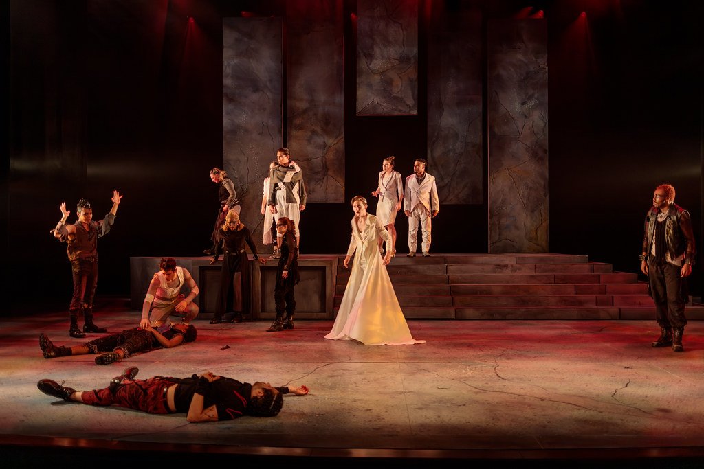 Stage scene from a theatrical performance with multiple actors, some standing on a platform and others lying or sitting on the stage floor, with a dark background and dramatic lighting.