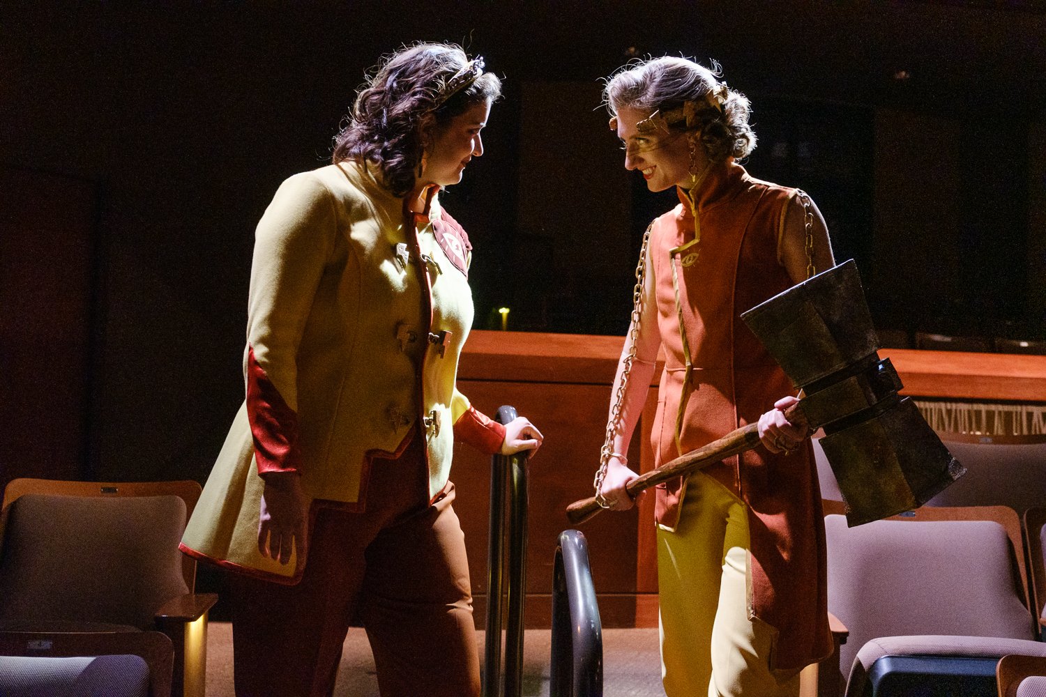 Two women dressed as Marvel's Thors in cosplay, standing face-to-face in a dark room with theater seats, one holding a globe-shaped prop and the other holding a large hammer.