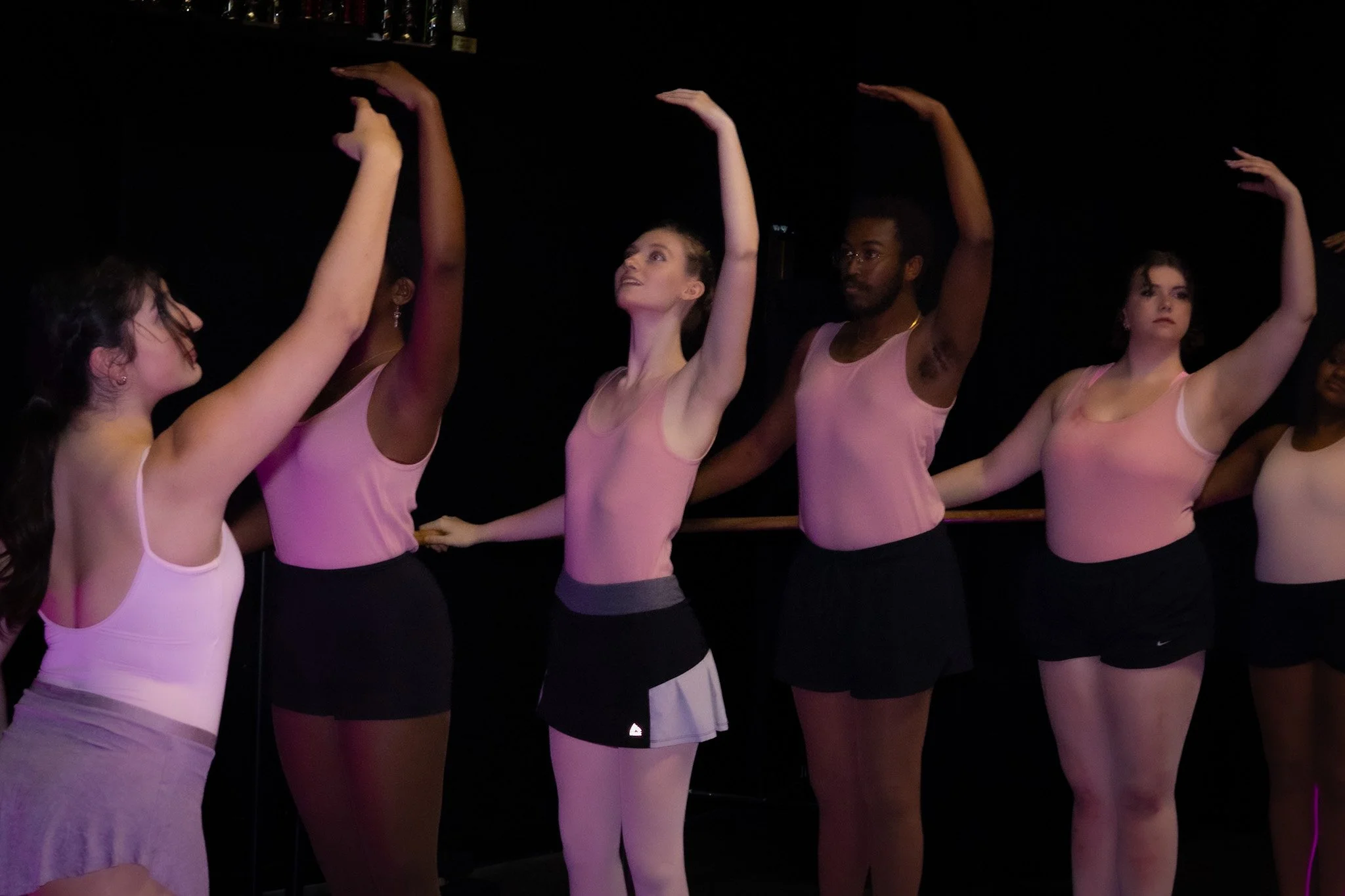 Five ballet dancers in pink leotards and black shorts practicing at ballet barre in a dark studio.