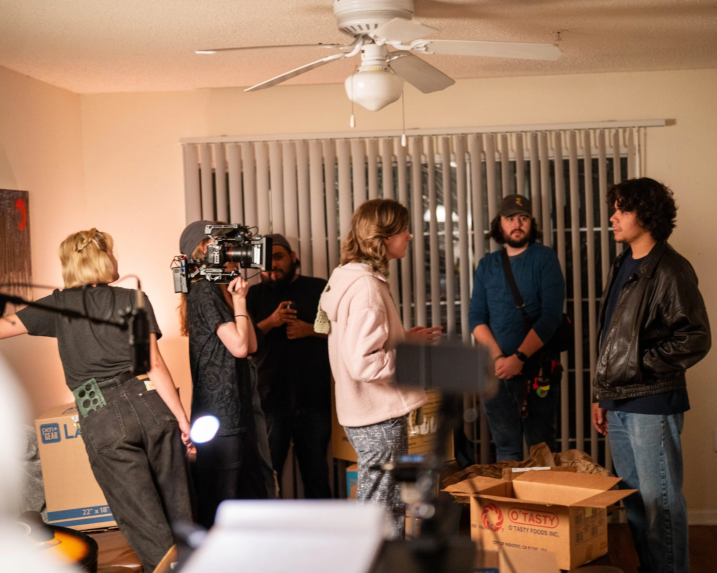 A group of people, including a woman with a camera and others talking, are inside an apartment with boxes on the floor and blinds on the window.