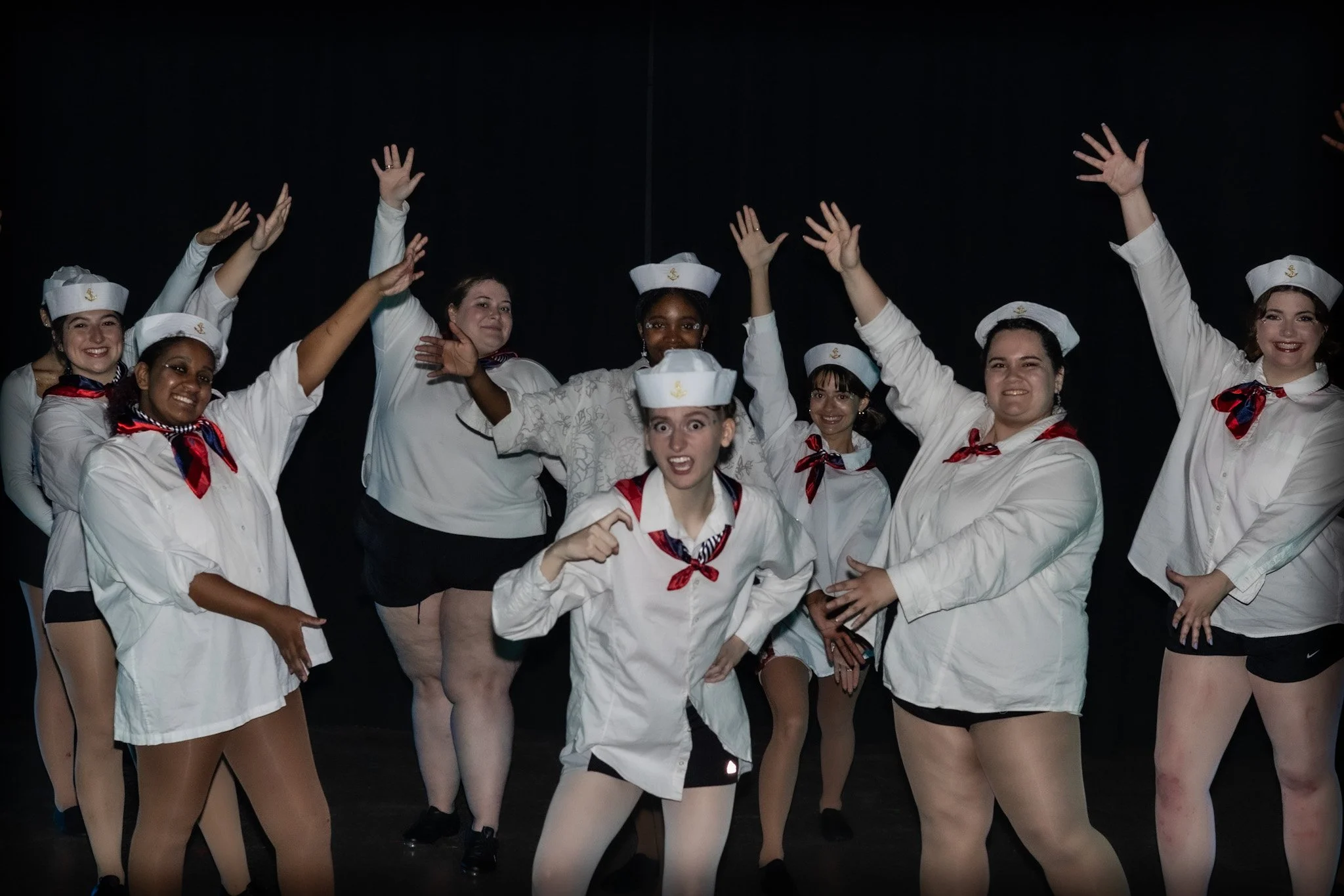 Group of diverse women dressed as sailors, posing with hands raised, on stage with a black background.