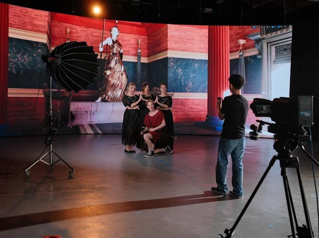 A video shoot in a studio with a large digital backdrop displaying a woman in a flowing dress. Five people pose in front of the backdrop, with one seated in a chair and four standing behind. A person in casual clothing records the scene with a smartp