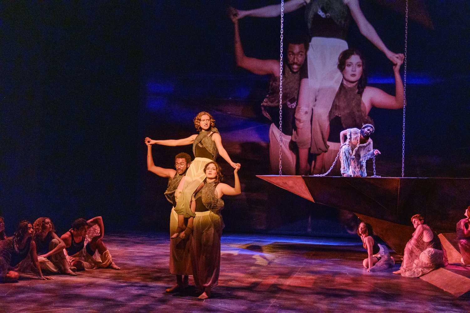 A theatrical stage performance with dancers, some forming a human pyramid, and others sitting or kneeling on the stage. Large screens in the background display images of two performers. The scene is lit with purple and orange lighting.